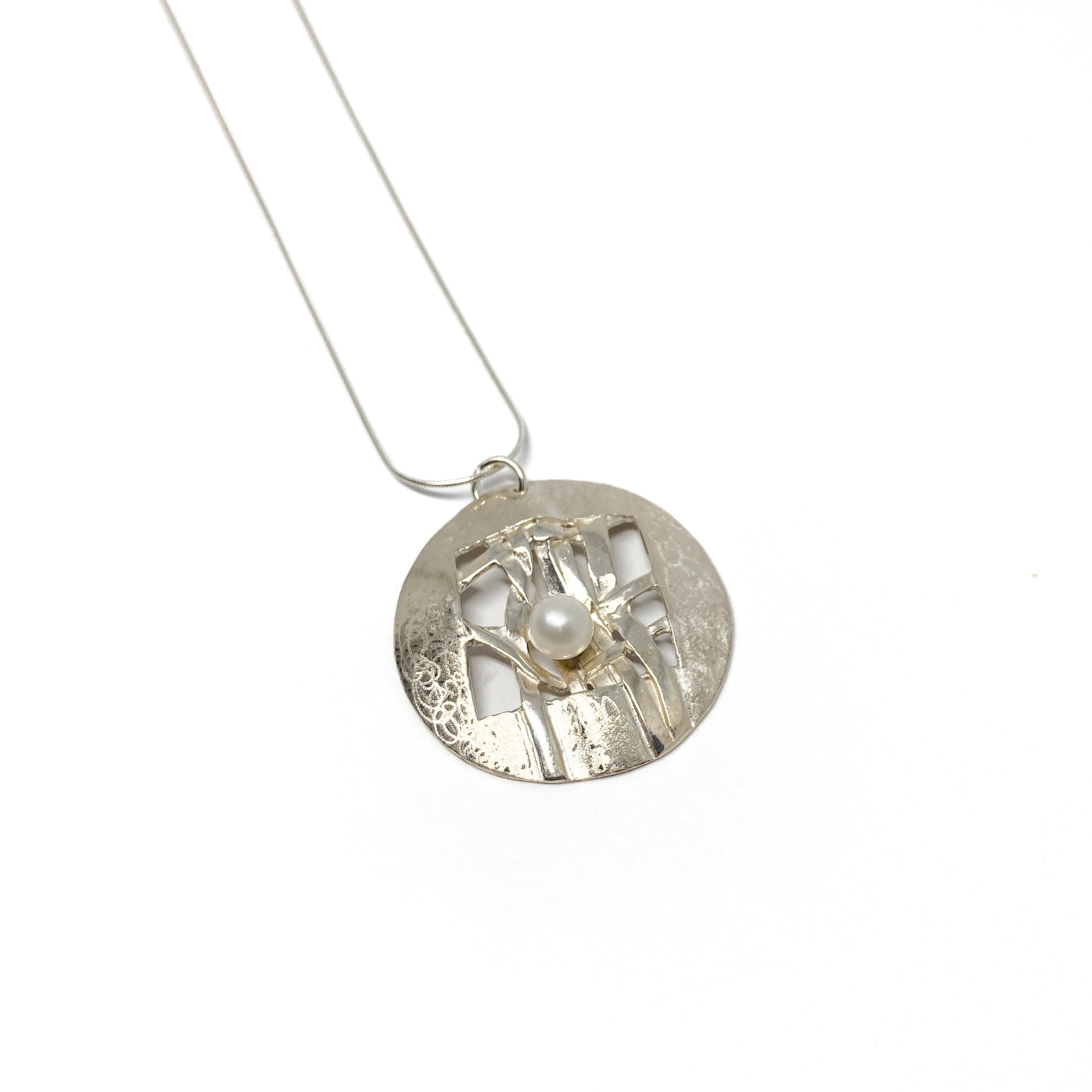 A silver pendant necklace with a circular disc pendant featuring a woven design and a pearl accent. The pendant hangs from a delicate silver chain.