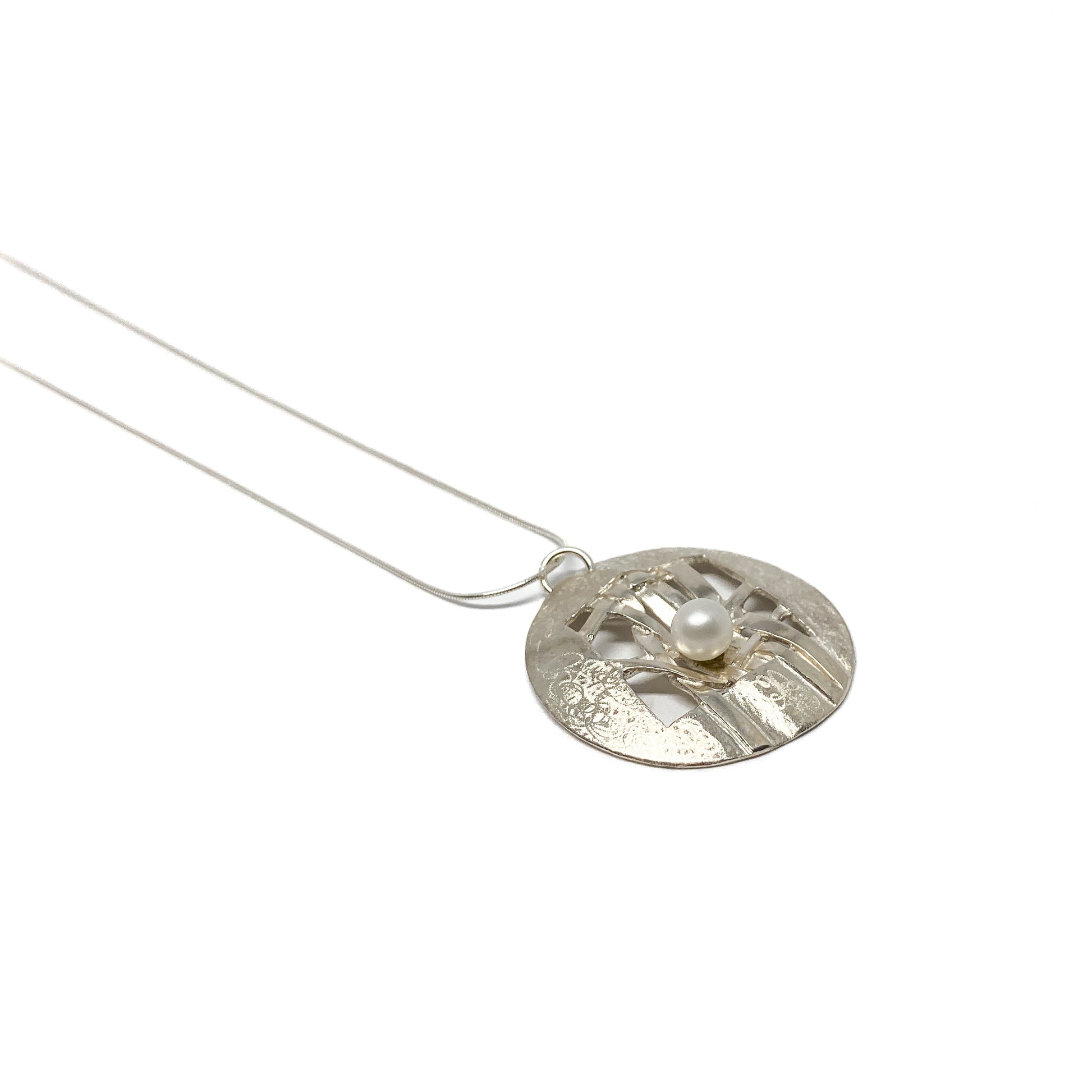 A silver pendant necklace with a circular disc pendant featuring a woven design and a pearl accent. The pendant hangs from a delicate silver chain.