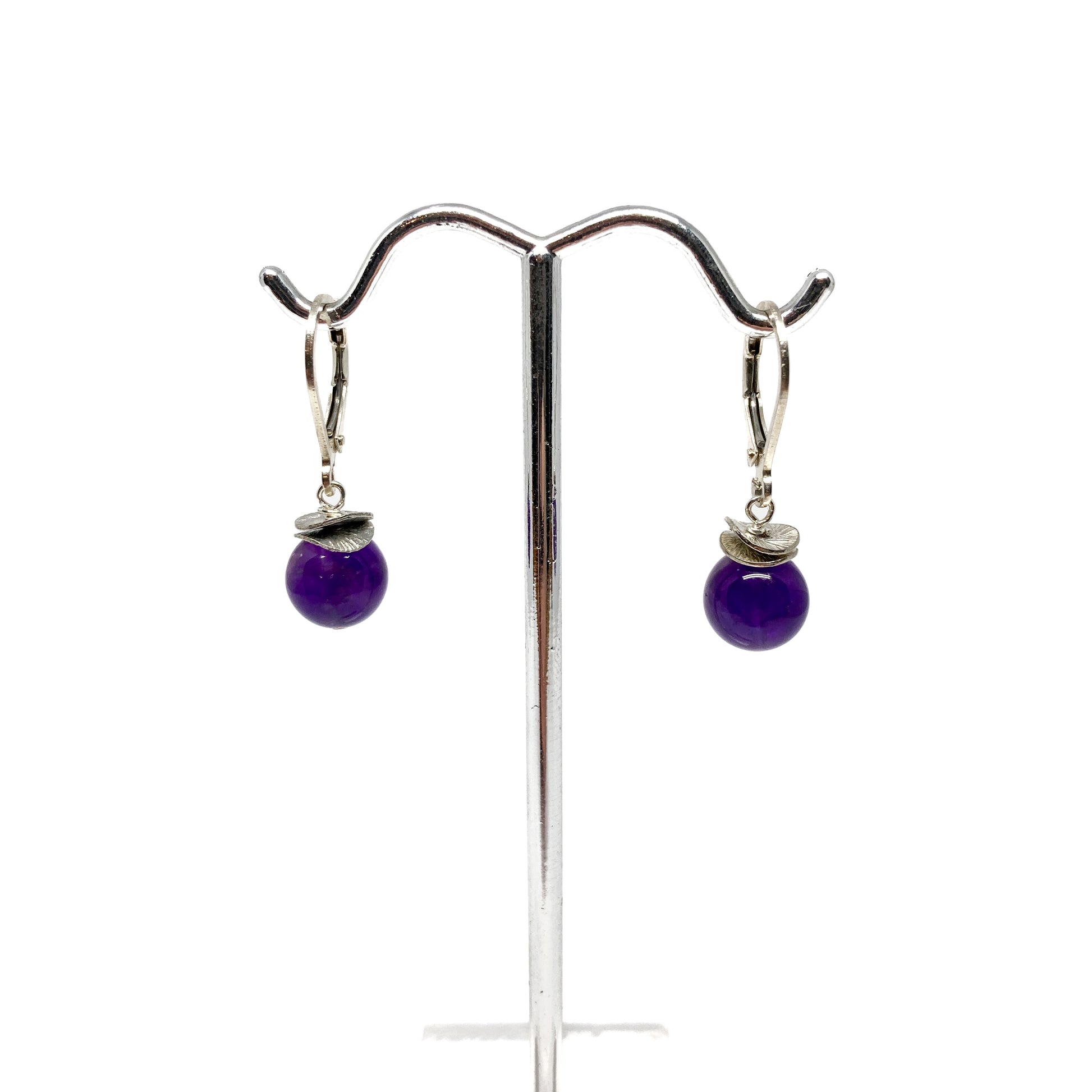 A pair of silver lever-back earrings with purple beads and a textured, petal-like design, displayed on a metal earring stand. The earrings have a secure lever closure.