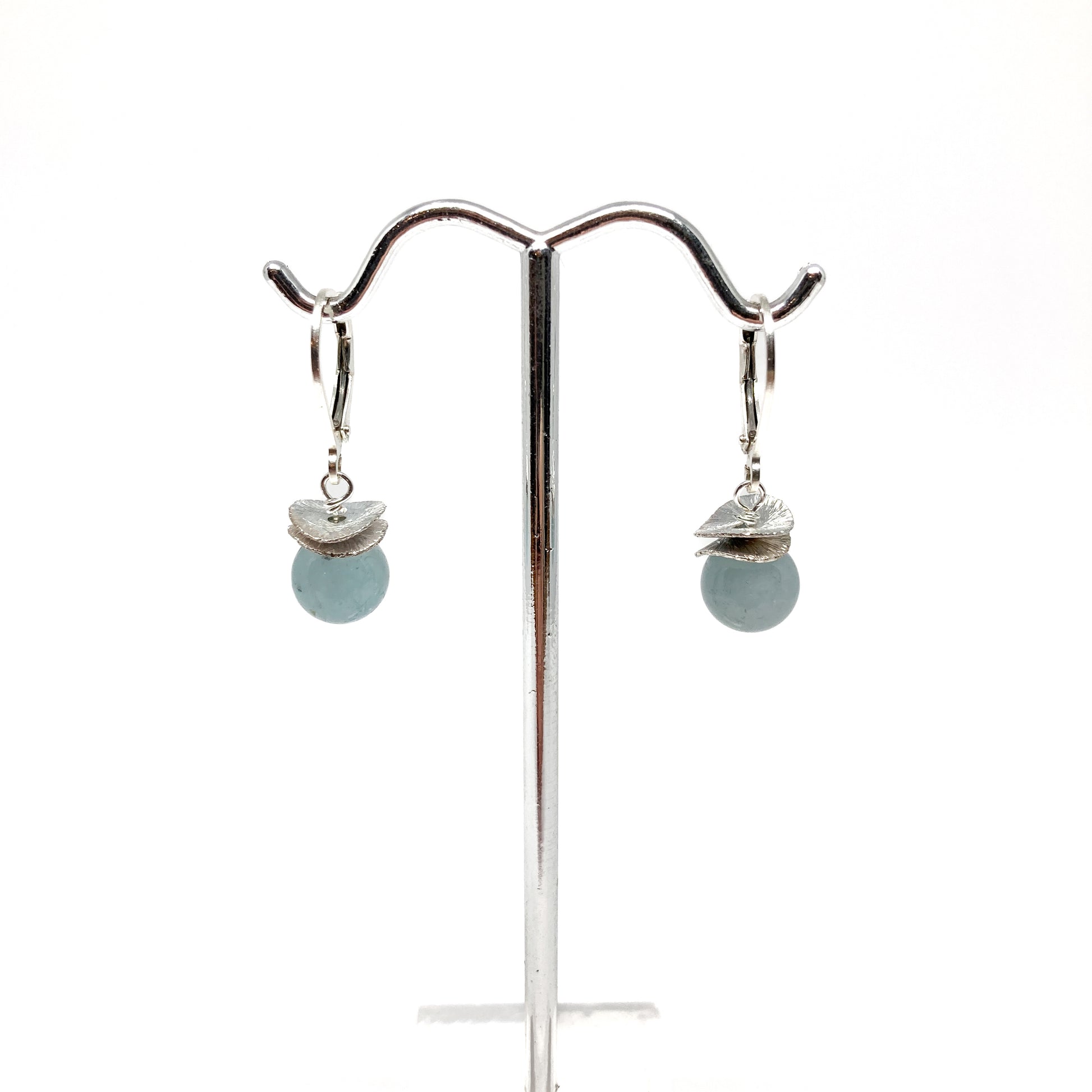 A pair of silver lever-back earrings with light blue beads and a textured, petal-like design, displayed on a metal earring stand. The earrings have a secure lever closure.