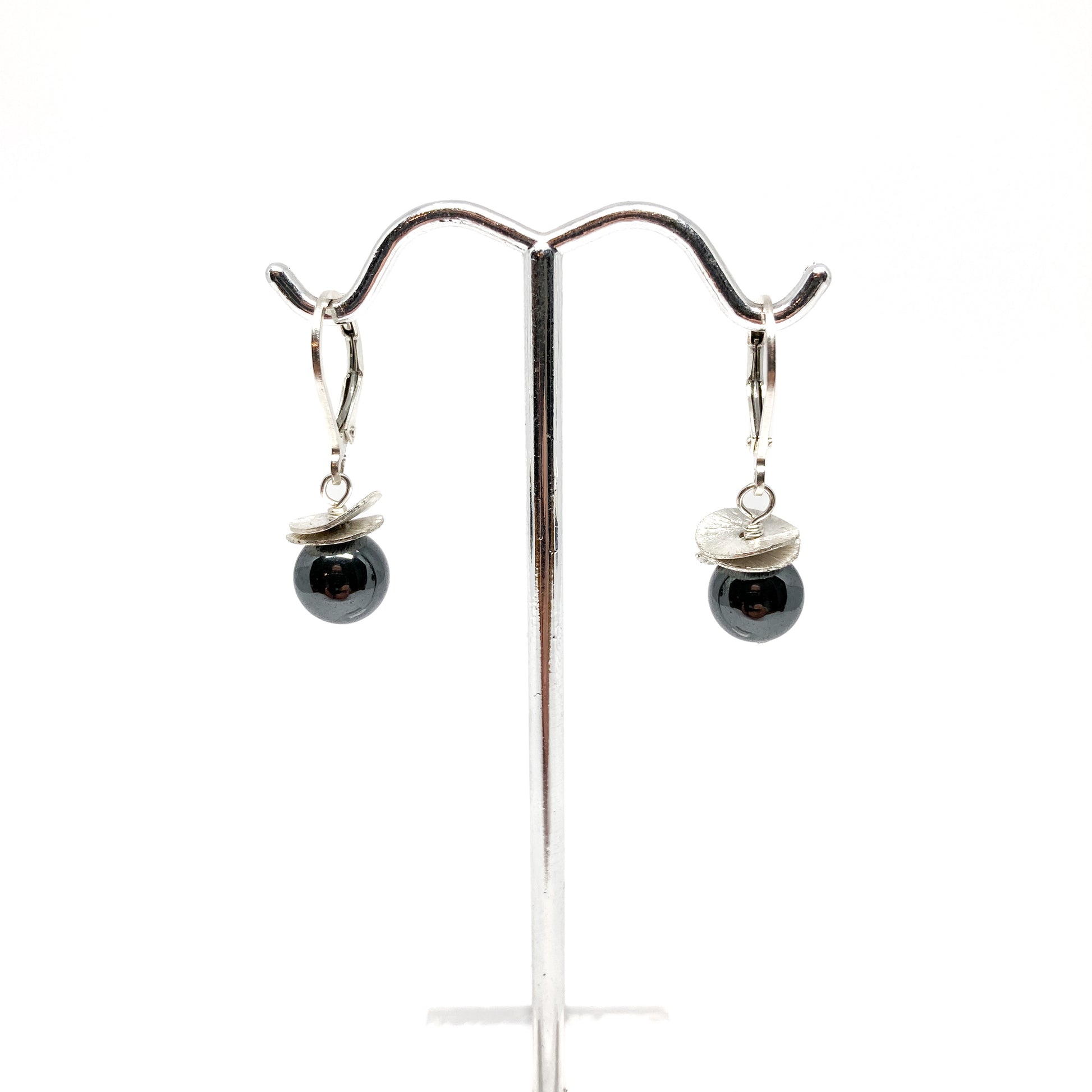 A pair of silver lever-back earrings with dark beads and a textured, petal-like design, displayed on a metal earring stand. The earrings have a secure lever closure.