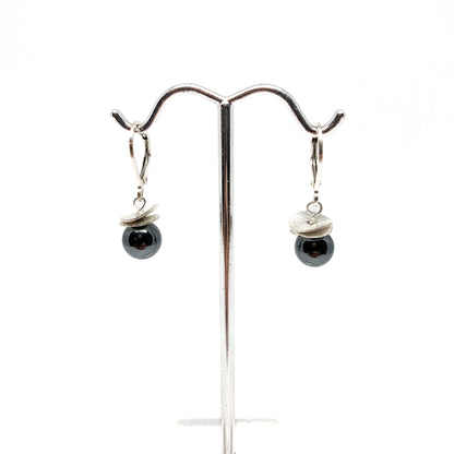 A pair of silver lever-back earrings with dark beads and a textured, petal-like design, displayed on a metal earring stand. The earrings have a secure lever closure.