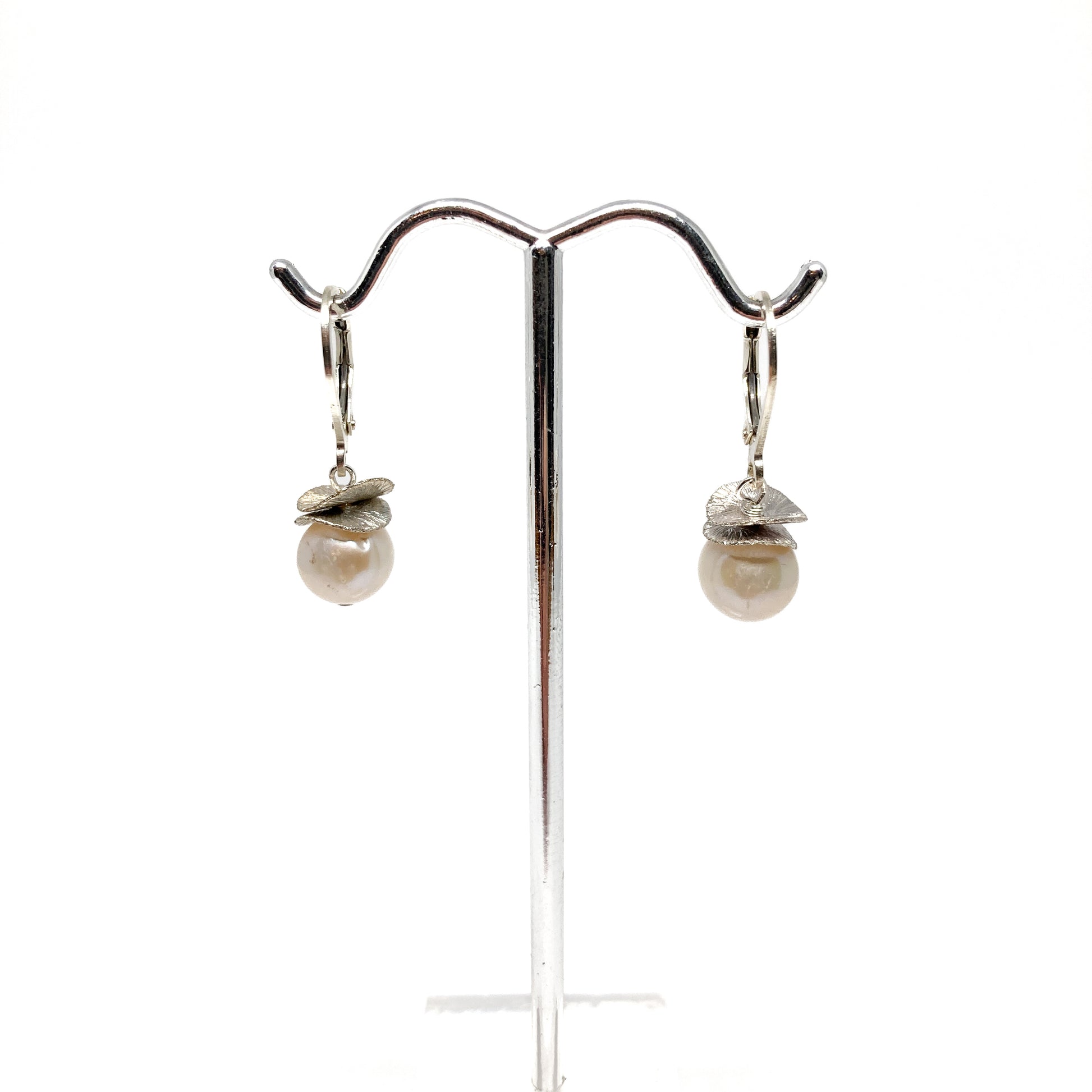 A pair of silver lever-back earrings with pearl beads and a textured, petal-like design, displayed on a metal earring stand. The earrings have a secure lever closure.
