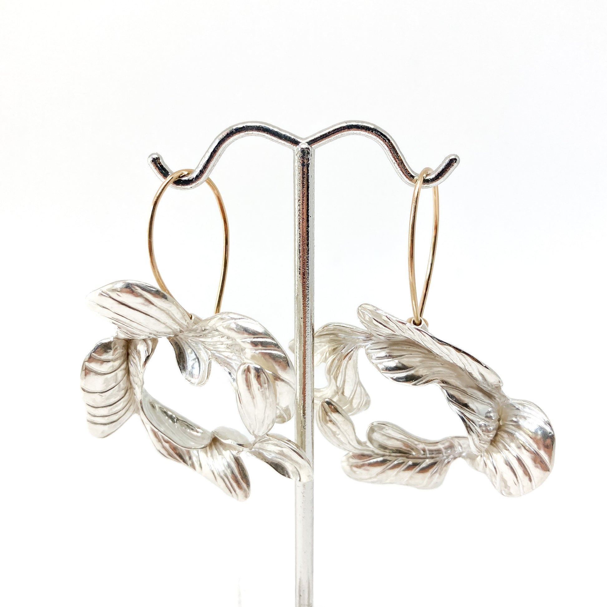 Closeness Earrings by Erin Christensen on a display. Silver with textured, abstract floral or leaf shapes and gold-toned hoops.