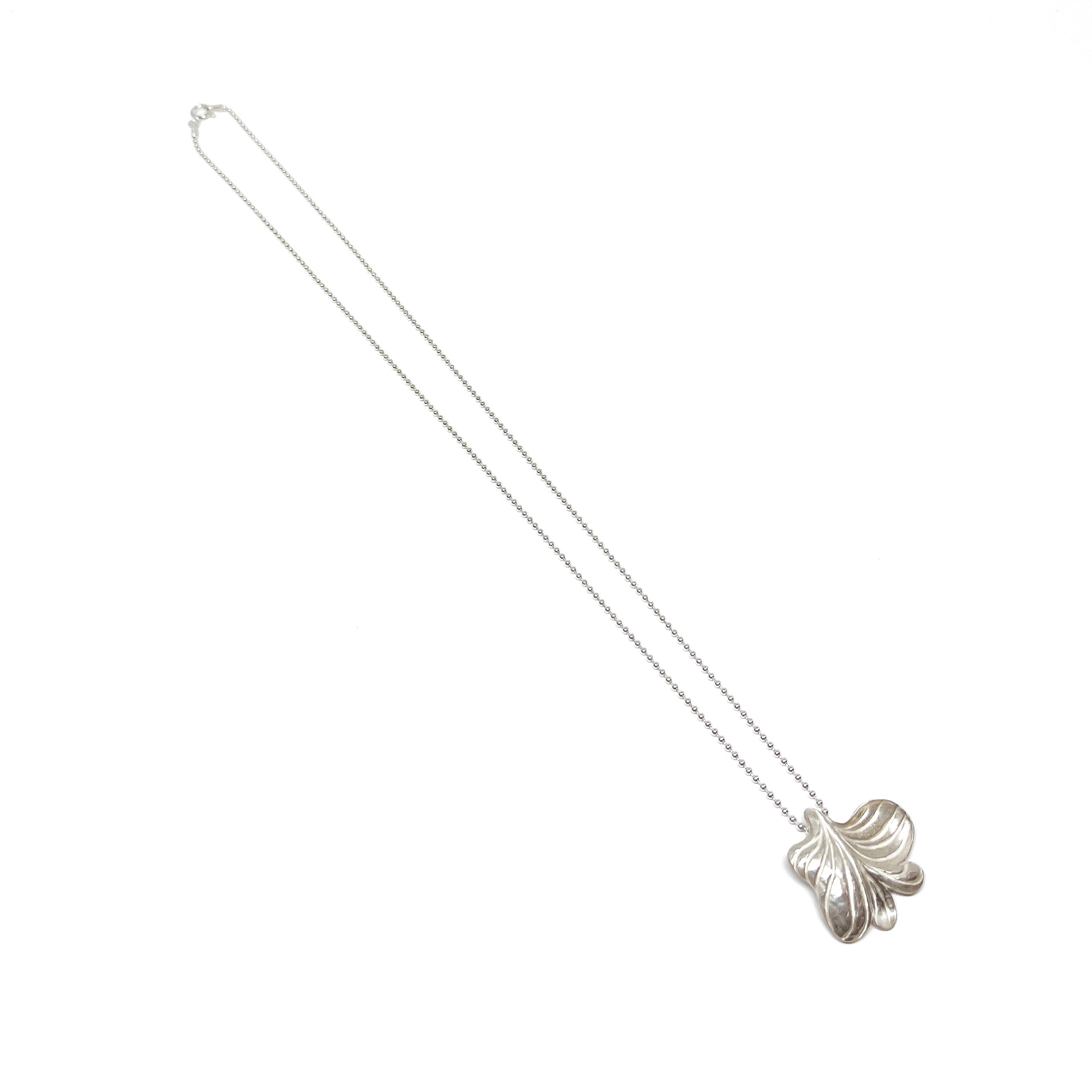 A long silver necklace featuring a large, textured pendant in an abstract floral or leaf-like shape.