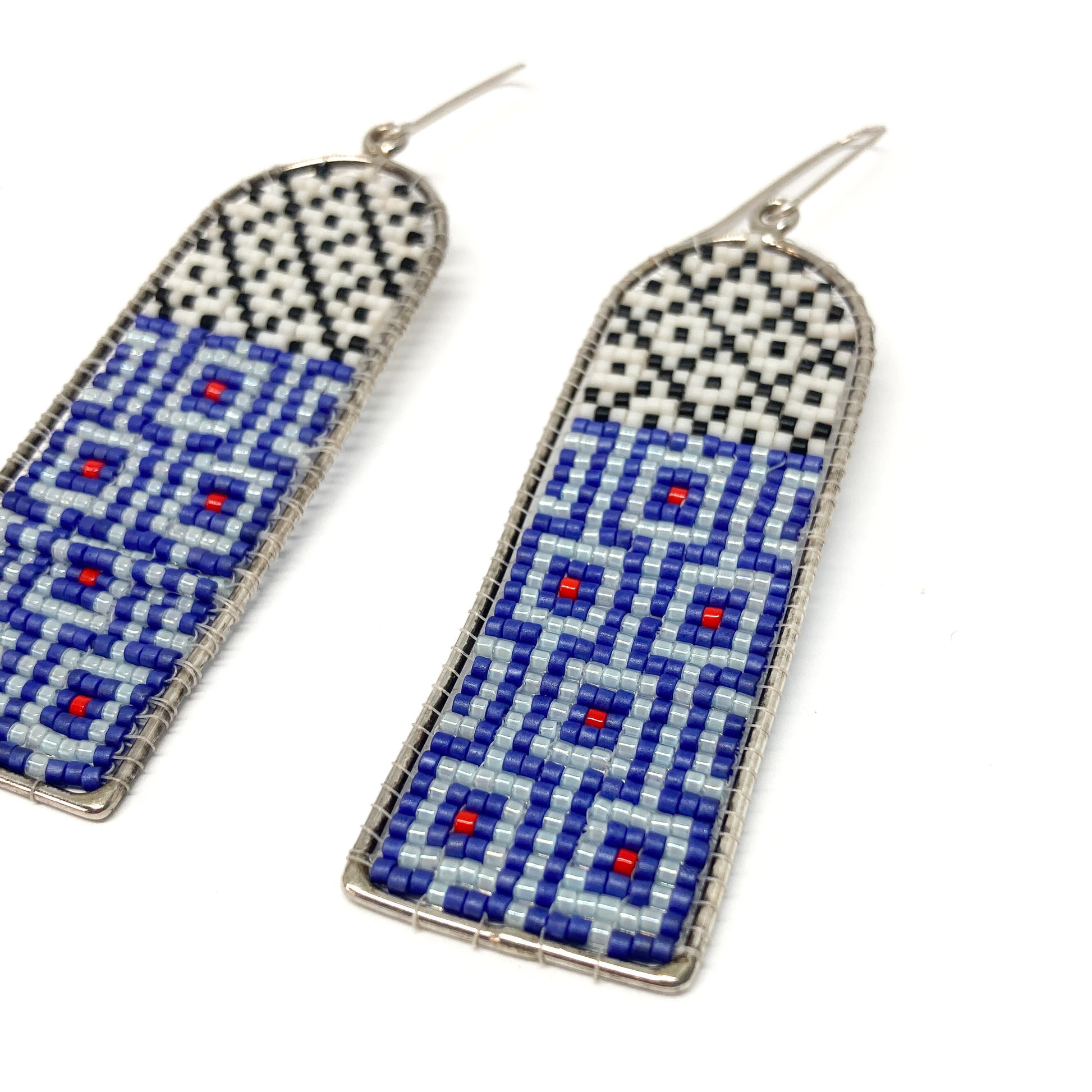 Two long, arched dangle earrings showcase detailed beadwork in white, black, blue, and red within silver frames.