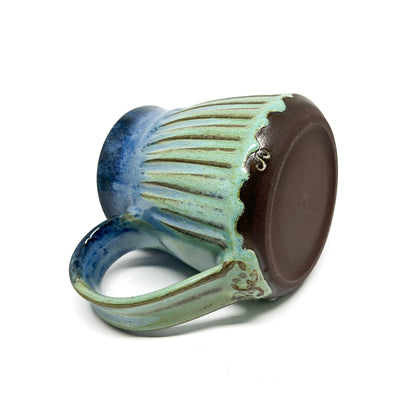 A close-up of the bottom of a ceramic mug with a ridged texture, featuring a blue-to-green gradient and a small inscription.
