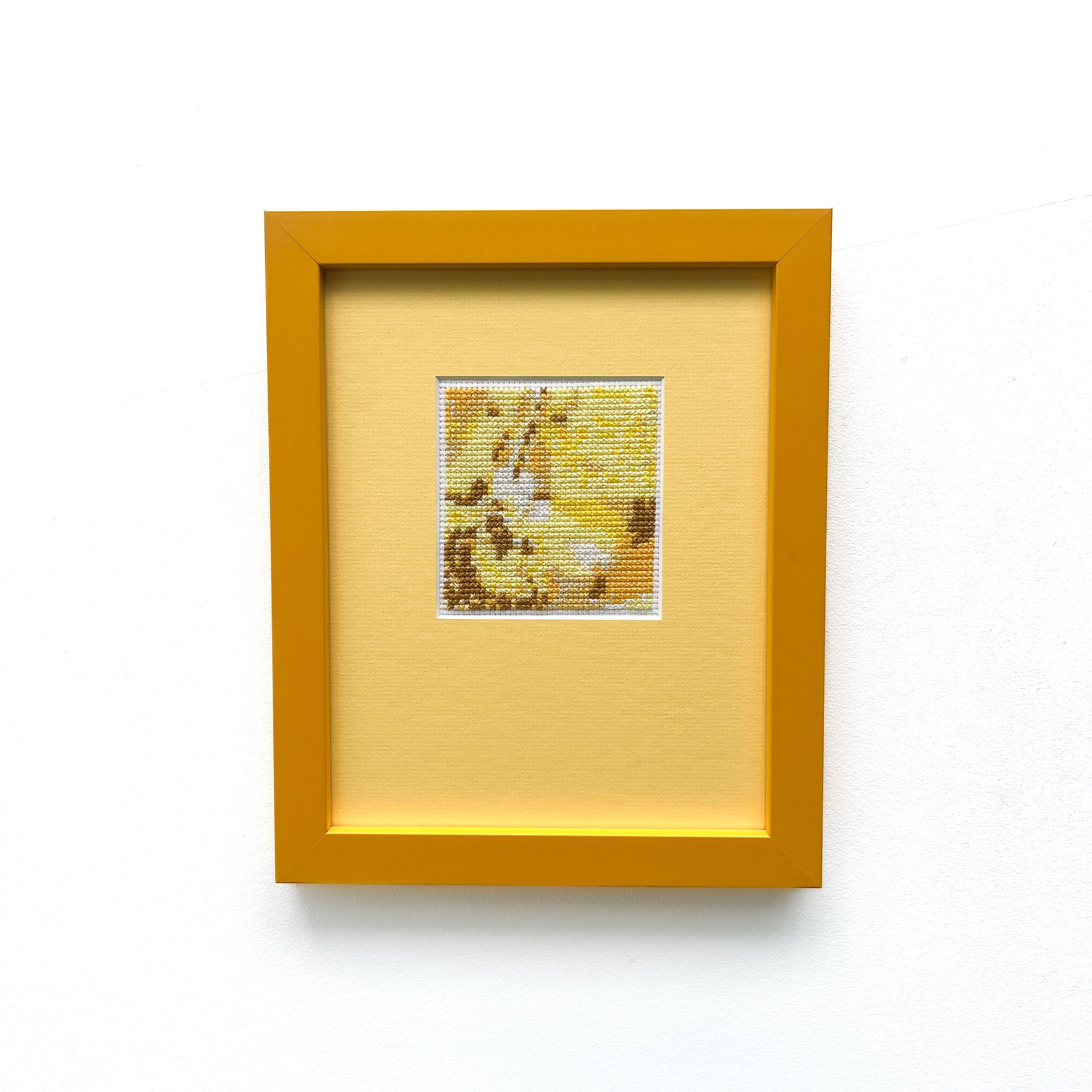 Framed "Yellow Monochrome" cross-stitch. A small square of stitched art is centered on a larger mat within a frame.