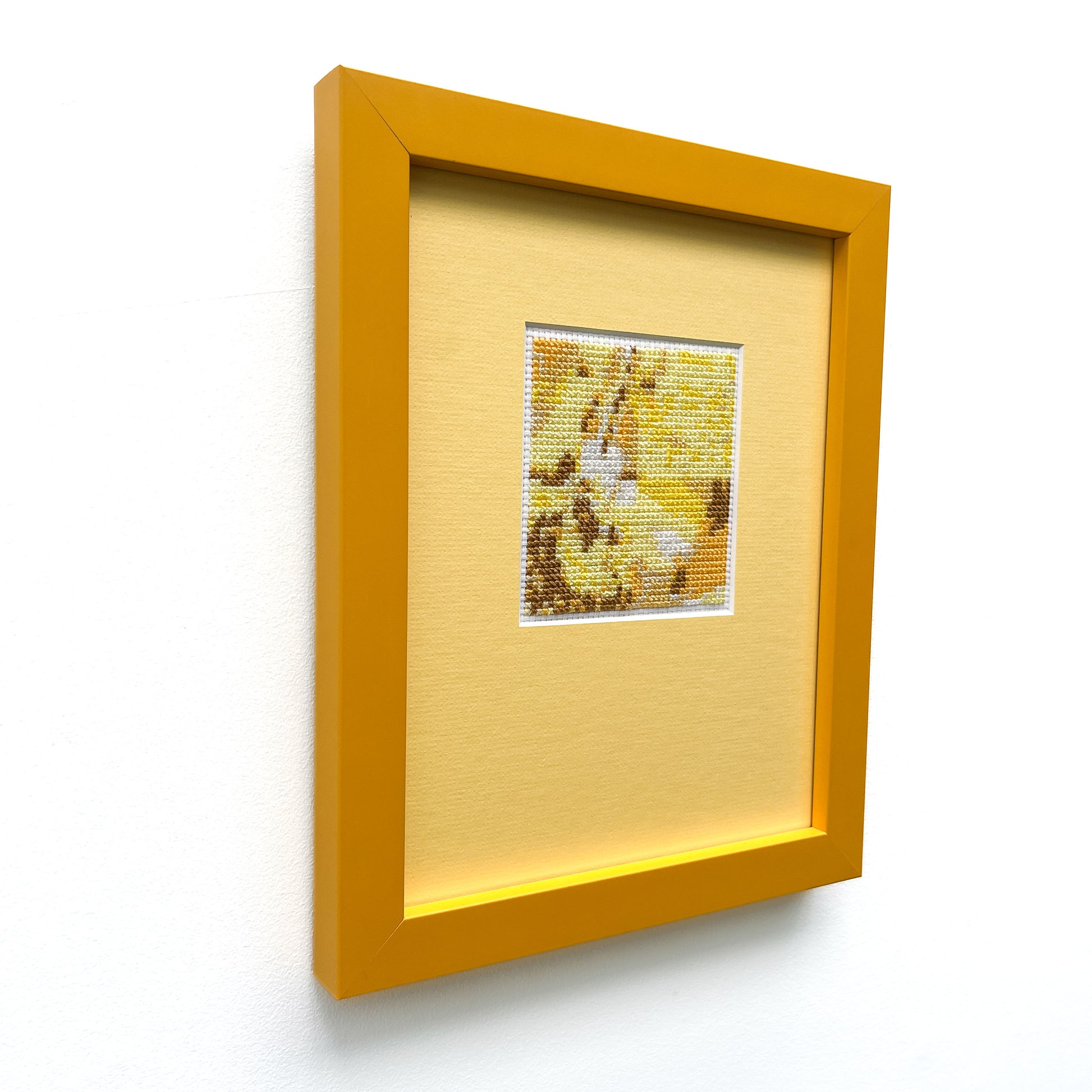 Framed "Yellow Monochrome" cross-stitch, viewed from the side. A small stitched square is centered on a mat within a frame.