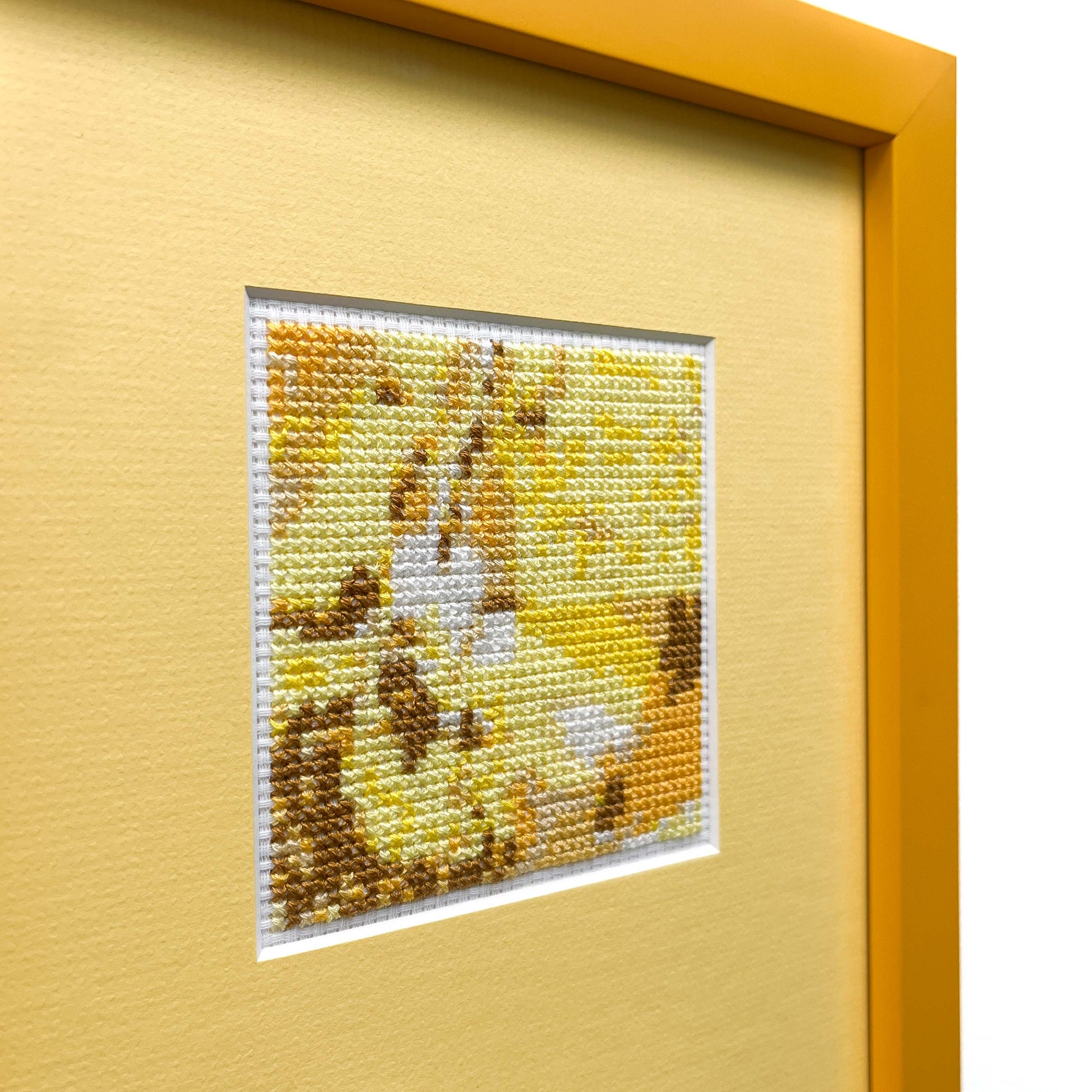 Close-up, side view of a framed "Yellow Monochrome" cross-stitch. The small stitched square is on a mat within a frame.