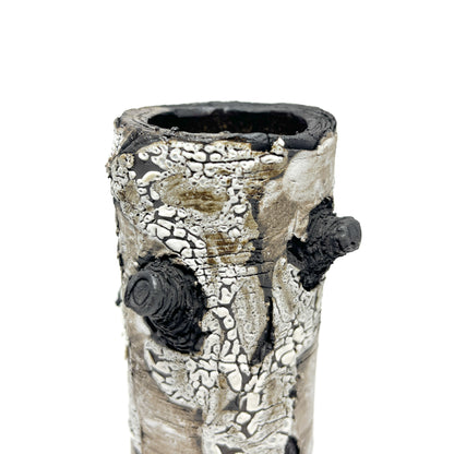 A close-up view of a cylindrical vase resembling birch bark with a textured surface, featuring black and white elements and a protruding branch-like detail.