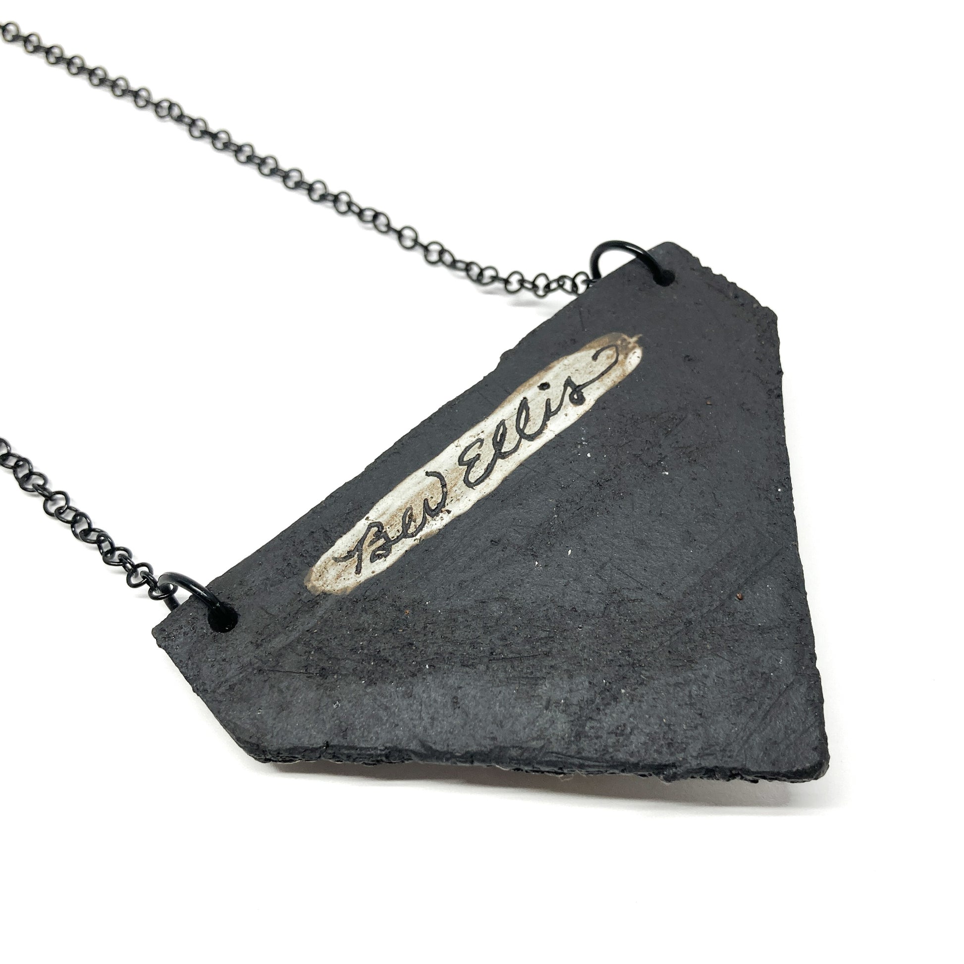 A triangular pendant necklace showing the artist's name "Bev Ellis" on the back, with a dark chain.