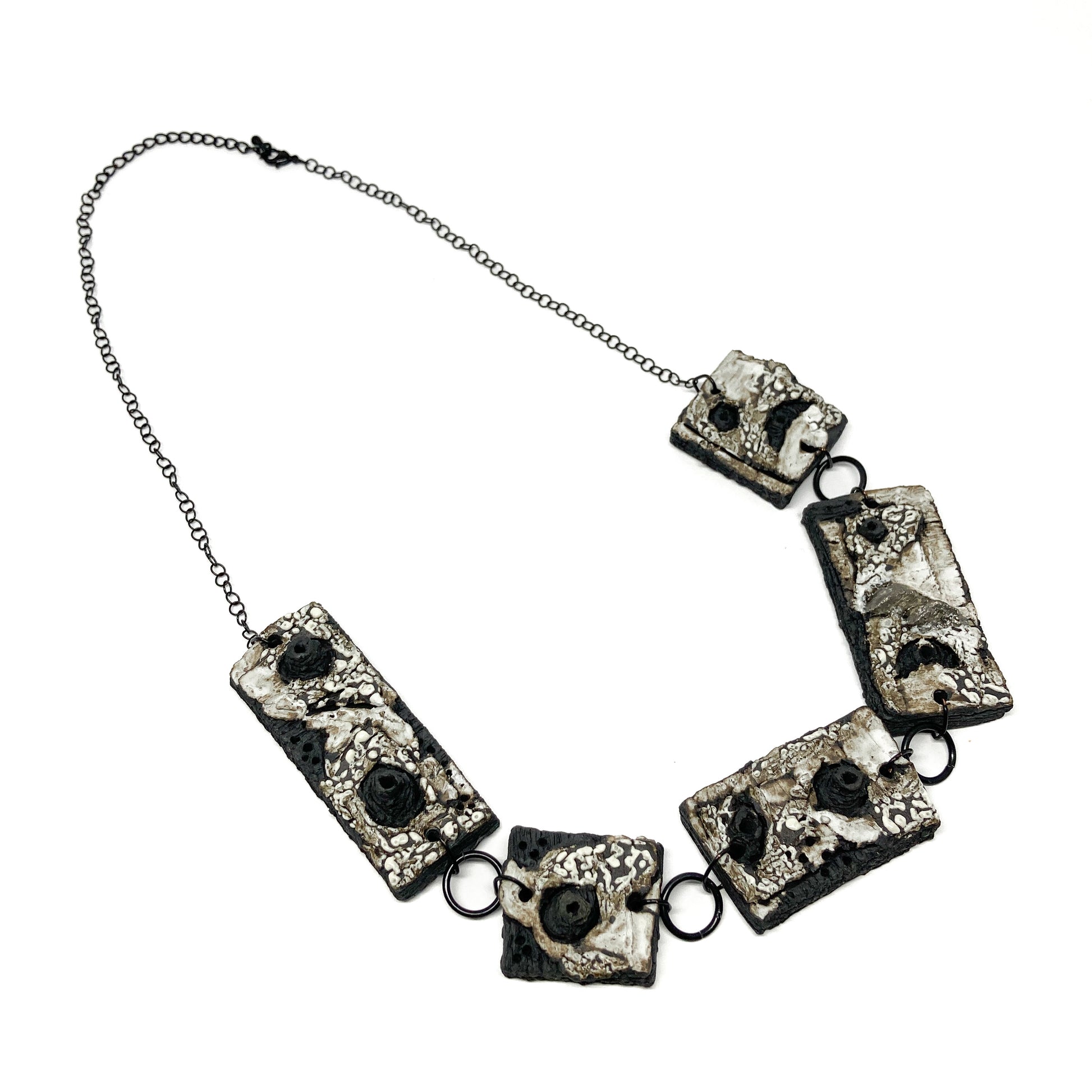 A necklace with five rectangular pendants featuring textured surfaces with black, white, and gray elements, linked by dark rings on a dark chain.