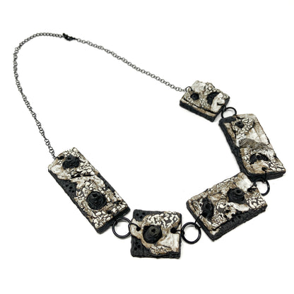 A necklace with five rectangular pendants, each featuring a textured surface with black, white, and gray elements, on a dark chain.