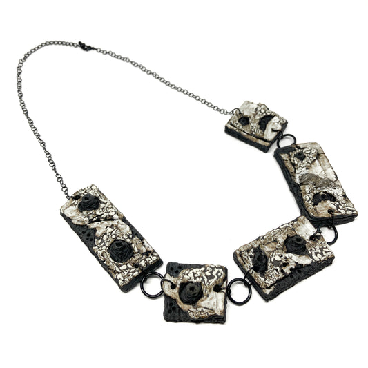 A necklace with five rectangular pendants, each featuring a textured surface with black, white, and gray elements, on a dark chain.