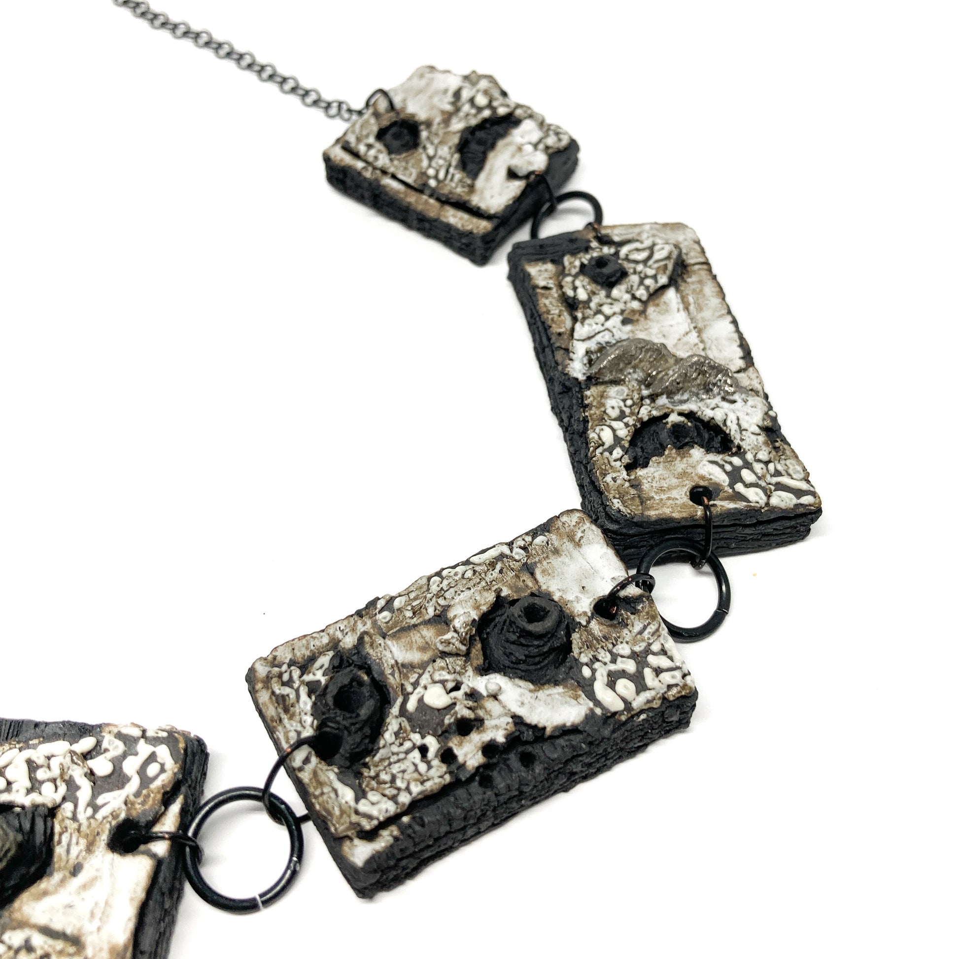 A close-up view of a necklace with rectangular pendants, featuring a textured surface with black, white, and gray elements, on a dark chain.