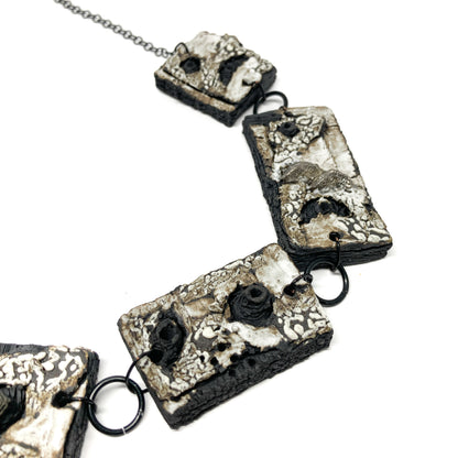 A close-up view of a necklace with rectangular pendants, featuring a textured surface with black, white, and gray elements, on a dark chain.
