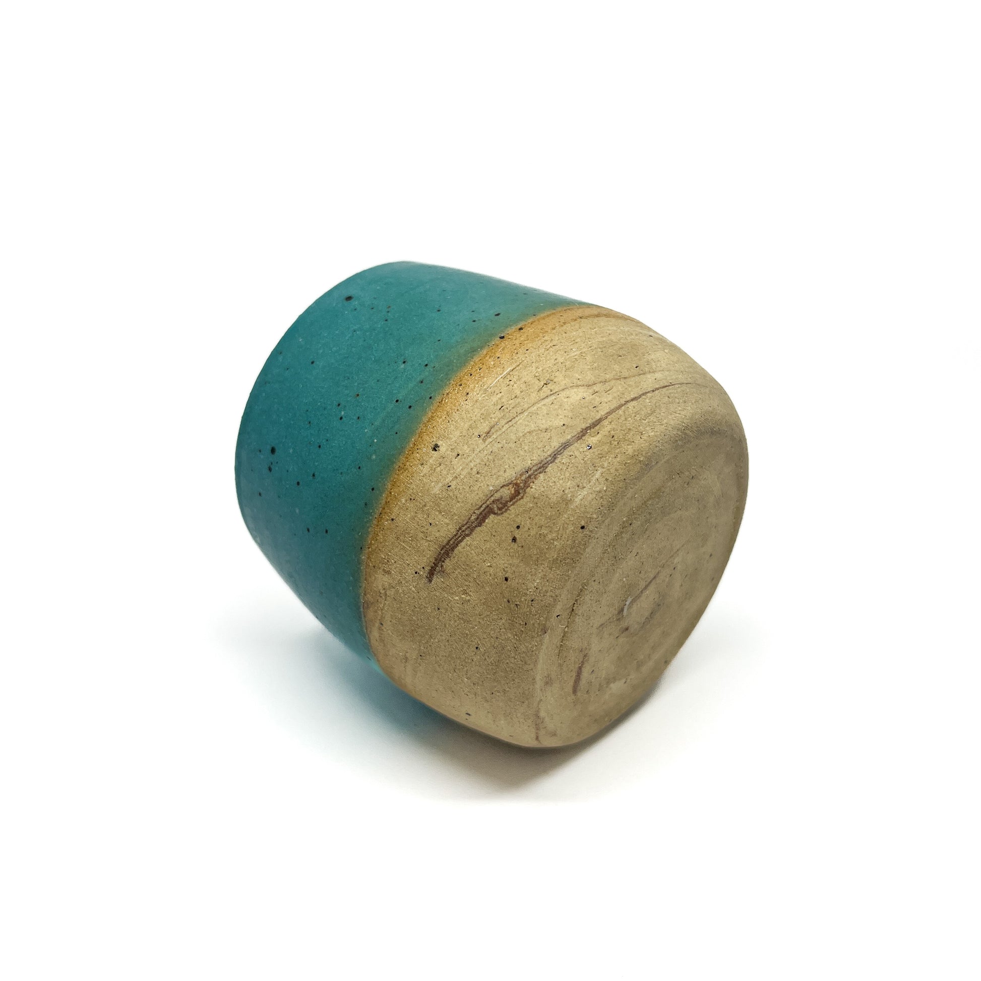 A small, cylindrical ceramic cup rests on its side, revealing a teal glazed top and a textured, unglazed base.
