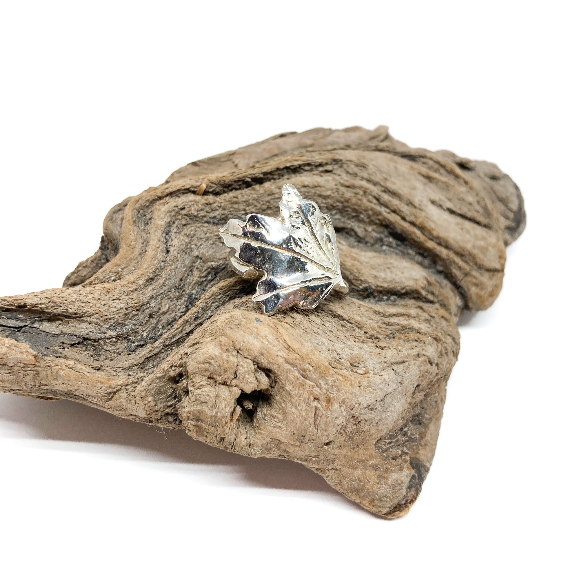A shiny silver ring shaped like a detailed maple leaf rests on a textured piece of driftwood.