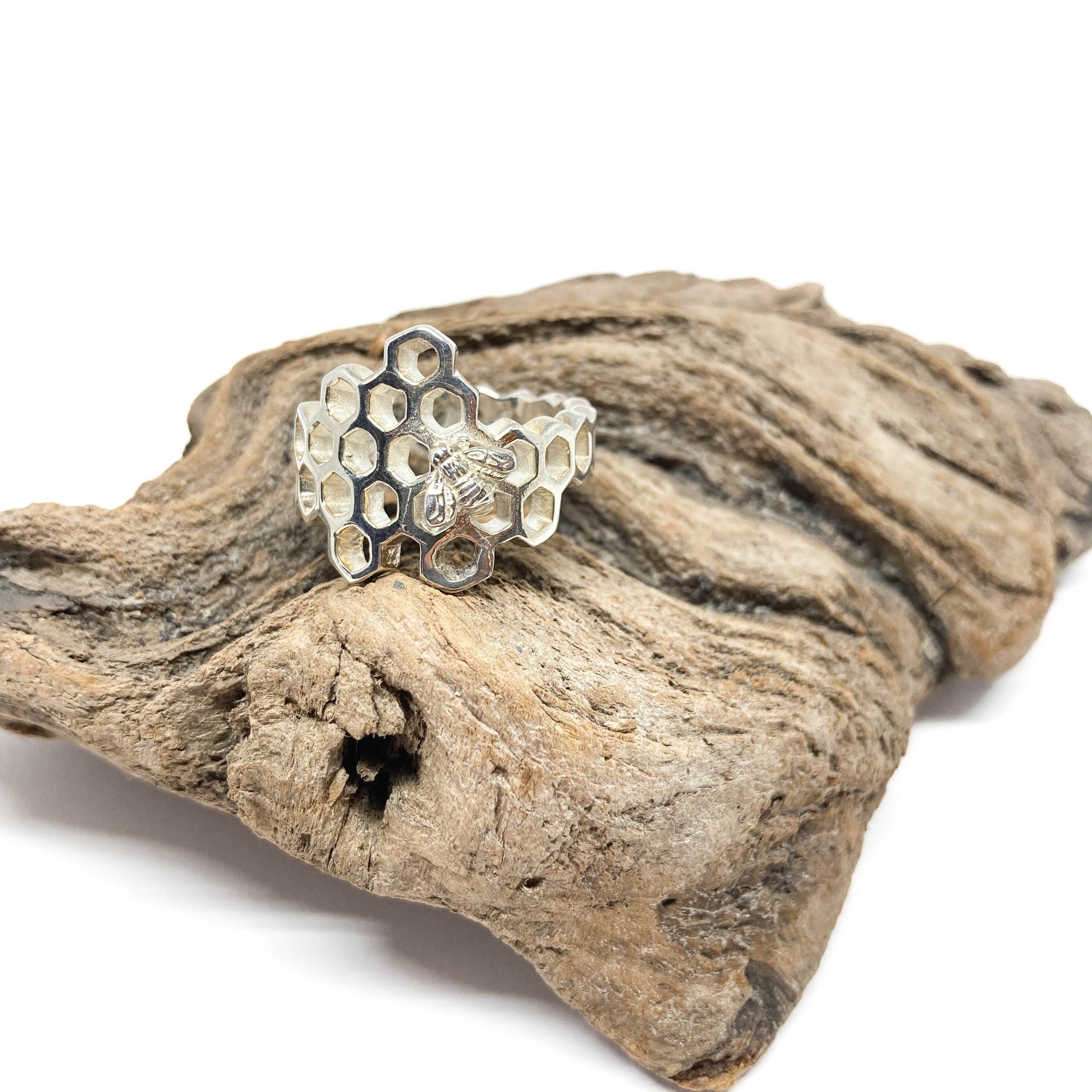 A shiny silver ring shaped like a honeycomb with a bee detail rests on a textured piece of driftwood.