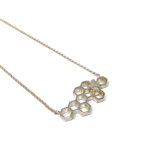 A delicate silver necklace features a horizontal honeycomb pendant with a tiny bee on one cell.