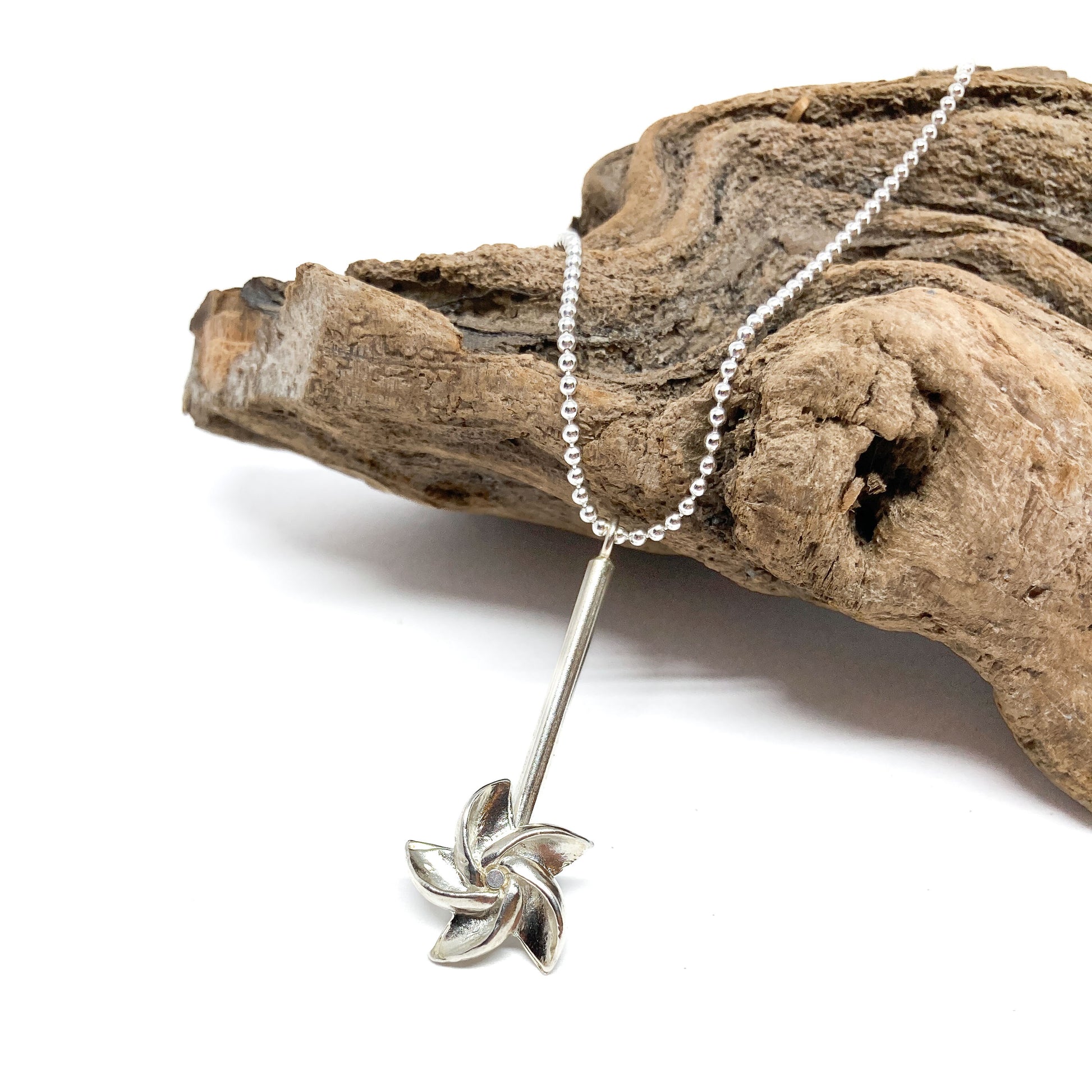 A silver ball chain necklace with a bar pendant and a pinwheel-like design rests on textured driftwood.