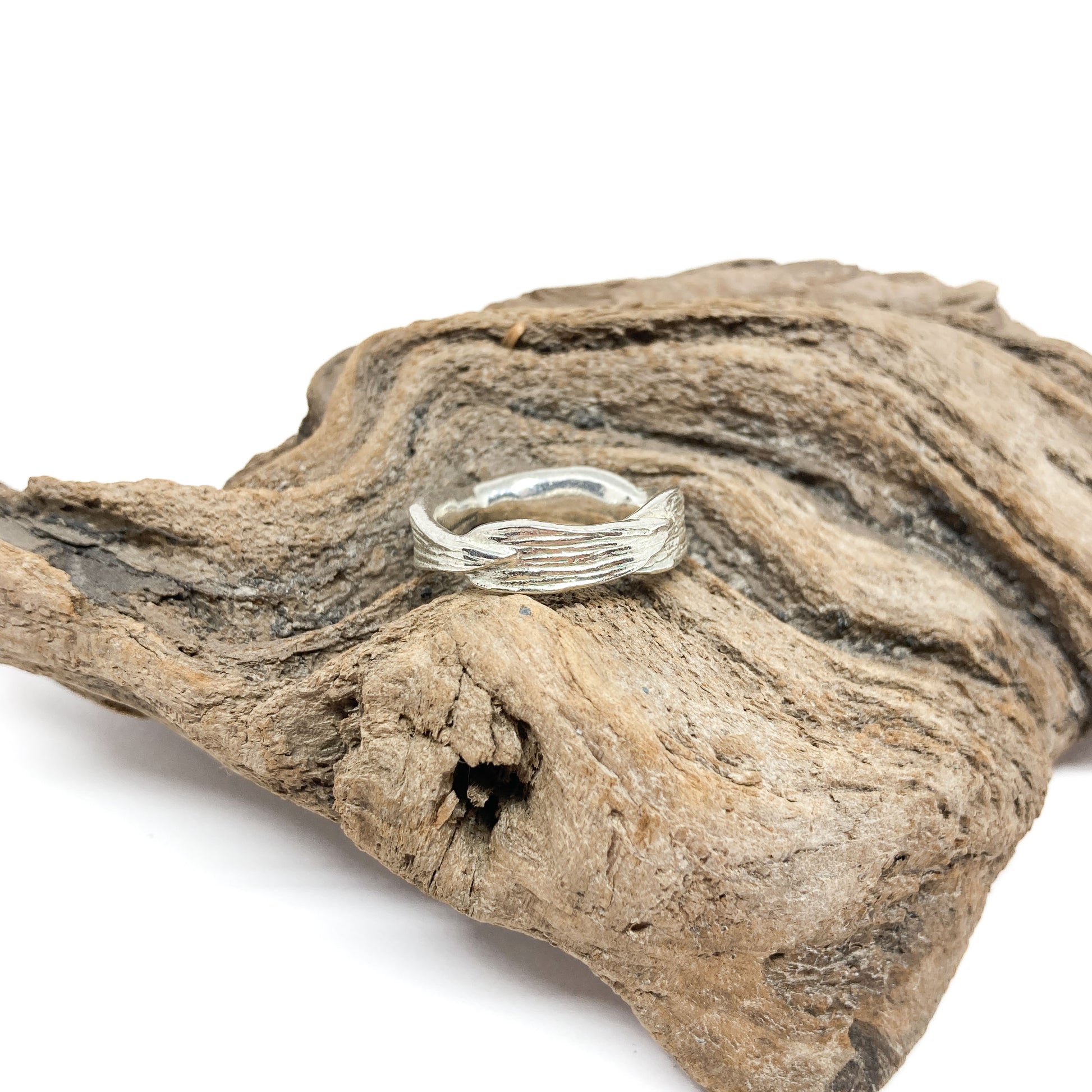 A shiny silver ring with a textured design resembling twisted bark rests on a piece of weathered driftwood.
