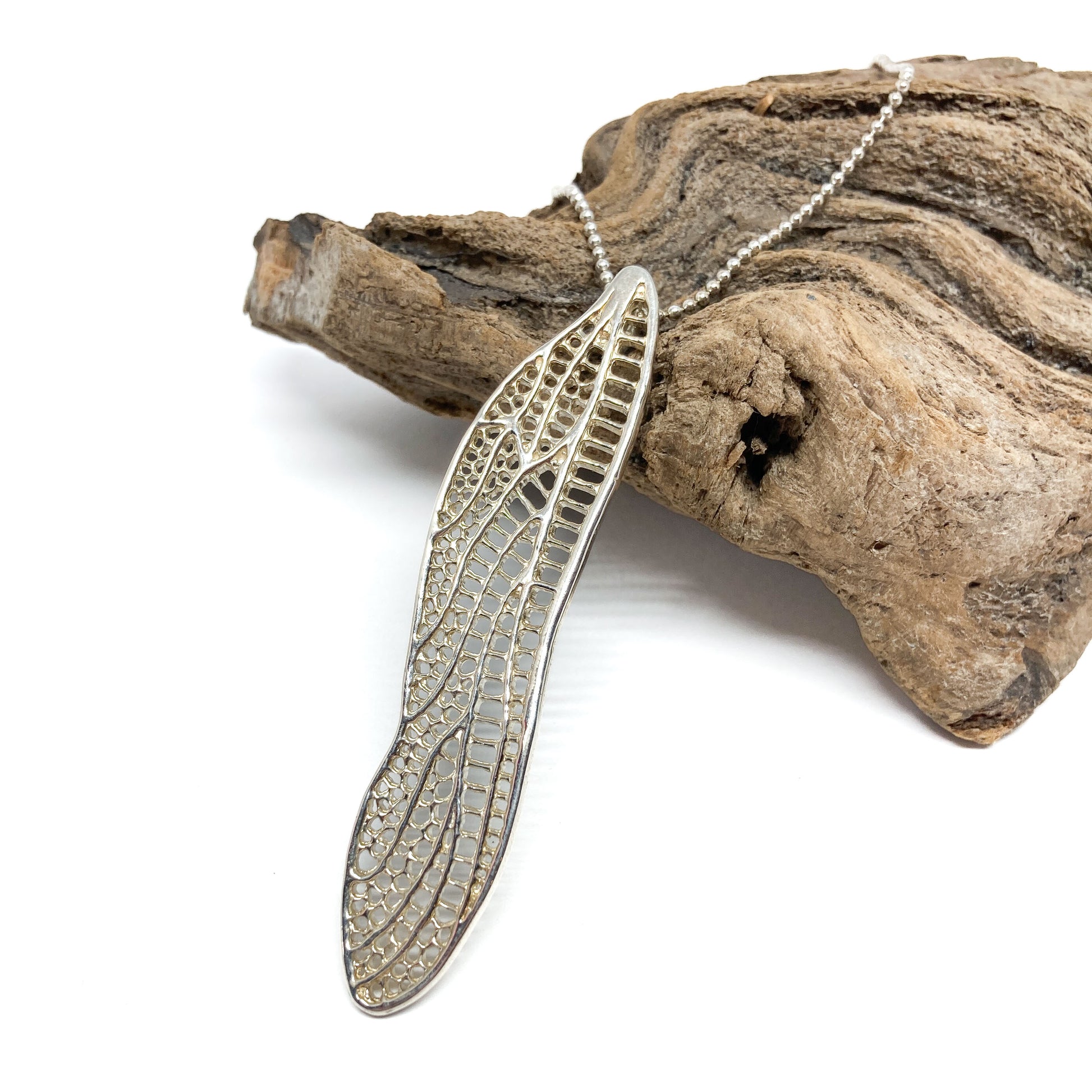 A silver necklace with a detailed dragonfly wing pendant rests diagonally on a textured piece of driftwood.