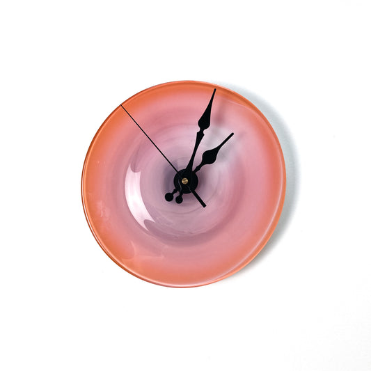 A round glass clock face with a gradient of pink, darker at the edges and lighter in the center, featuring ornate black hands.