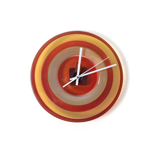 A round glass wall clock with concentric rings of red, yellow, and beige, featuring white hands and no numerals.