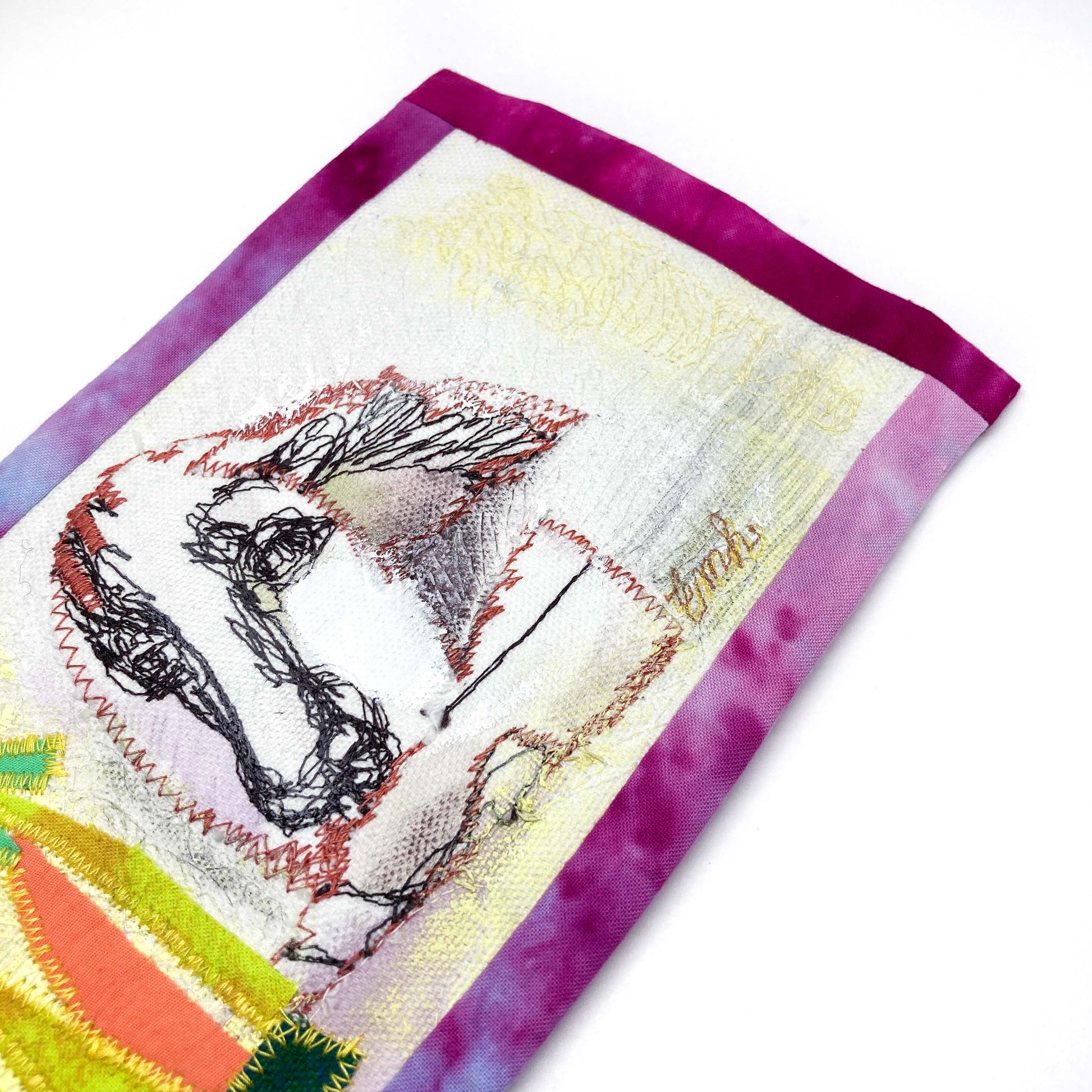 Close-up of a textile art panel showing a stylized line drawing of a dog's head and textured stitching on fabric with a dyed border.