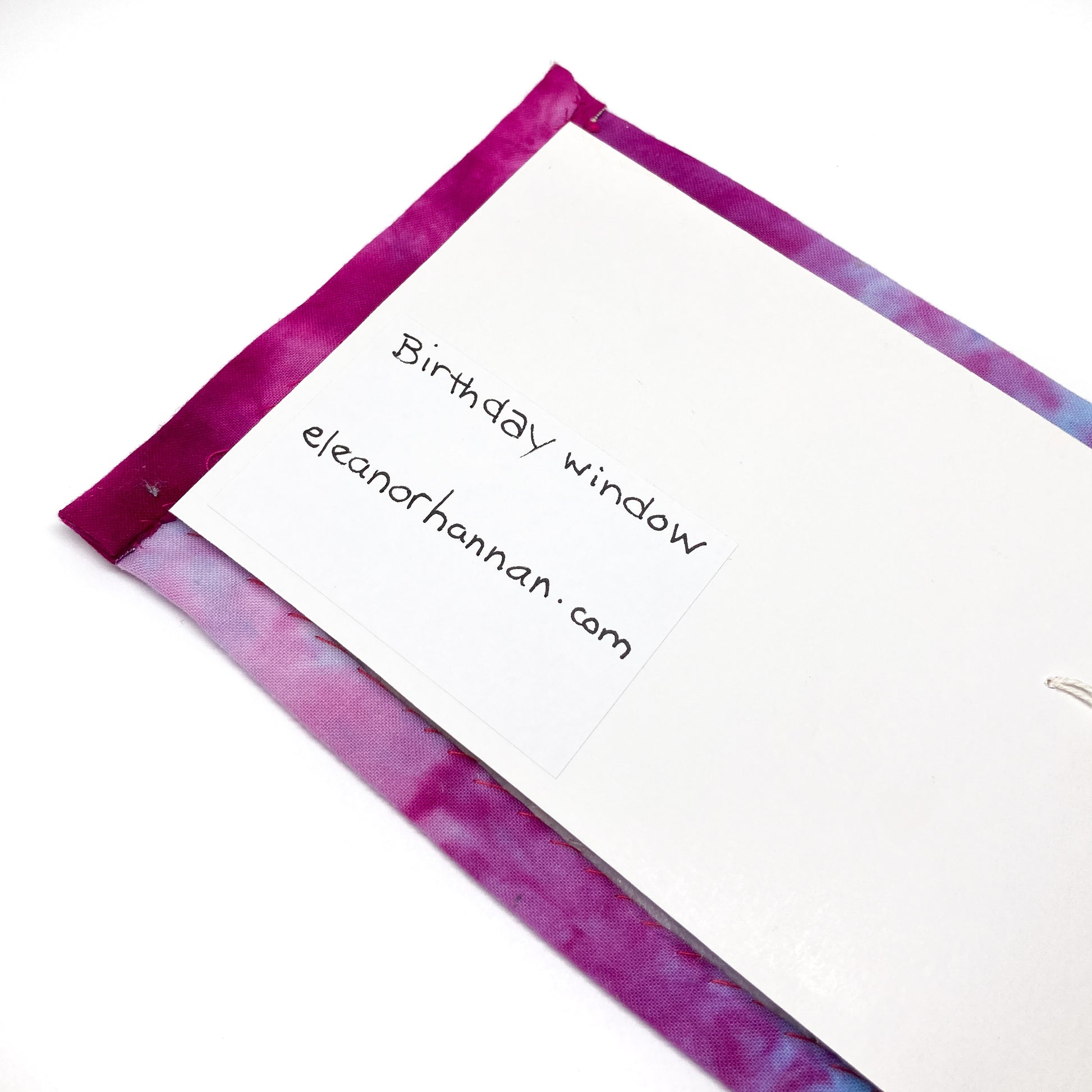 A white card with handwritten text "Birthday window" and "eleanorhannan.com" is pinned to a textile art piece with a dyed border.