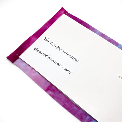 A white card with handwritten text "Birthday window" and "eleanorhannan.com" is pinned to a textile art piece with a dyed border.