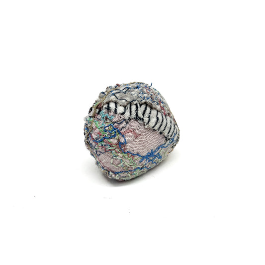 Embroidered Rock by Jane Kenyon: Round textured textile art resembling a rock, with layers of white, black, blue, and pink thread stitching.