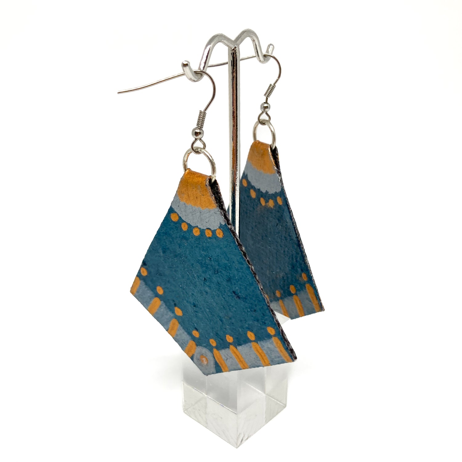 Dangling geometric earrings with blue, gold, and silver details hanging from a silver display stand with a clear base.