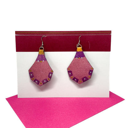 Two pink, purple, and gold geometric earrings with decorative edges hang from silver hooks on a display card.