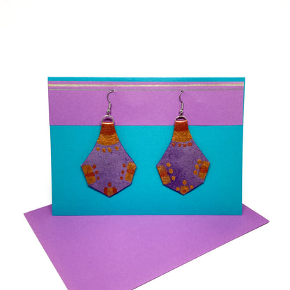 Two textured purple and gold geometric earrings with a unique shape hang from silver hooks on a display card.