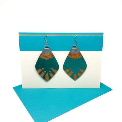 Two teal and copper geometric earrings with a leaf-like shape and radiating details hang from silver hooks on a display card.