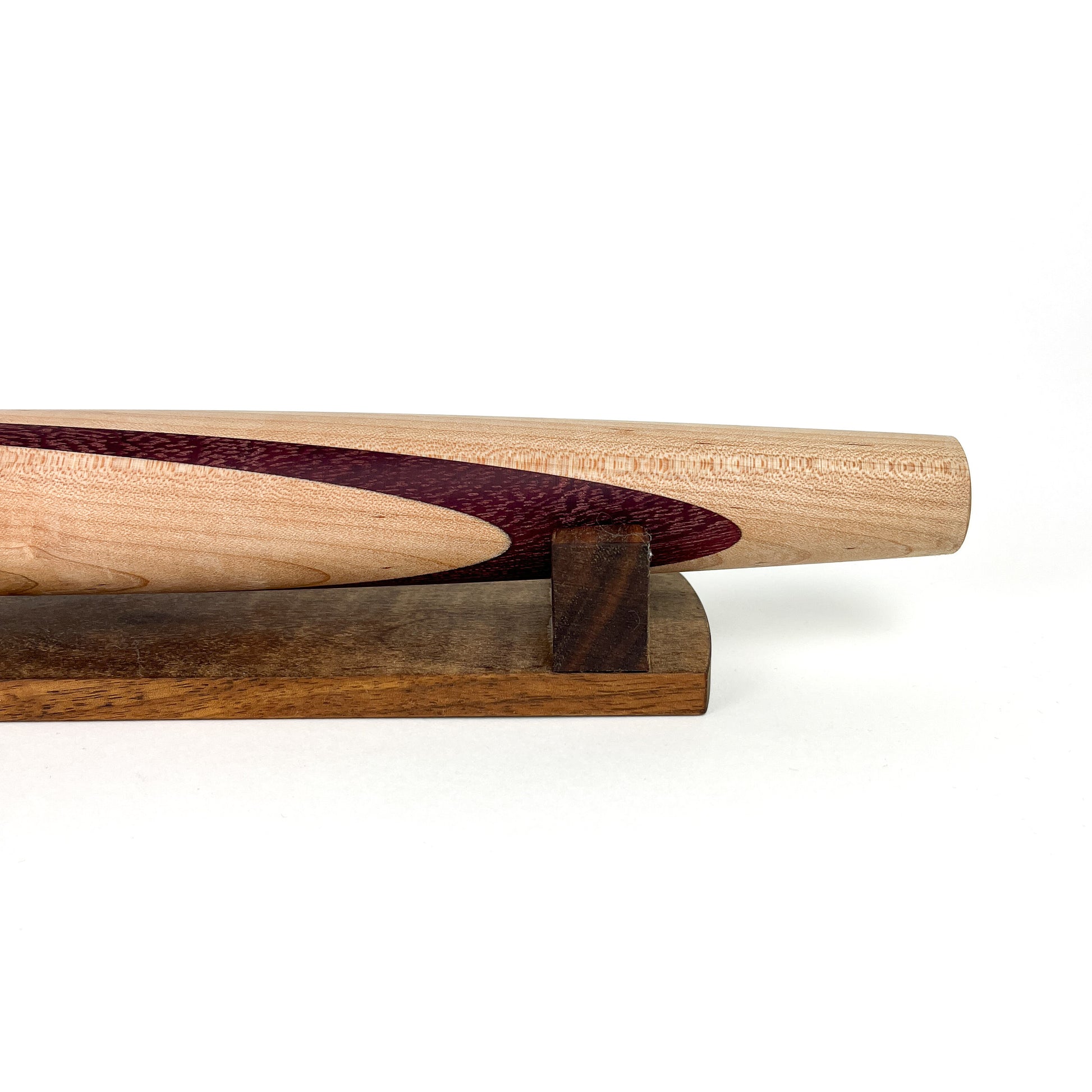 A close-up of a wooden French rolling pin with a dark purple inlay, resting on a dark wooden stand. The pin has a smooth, polished finish.