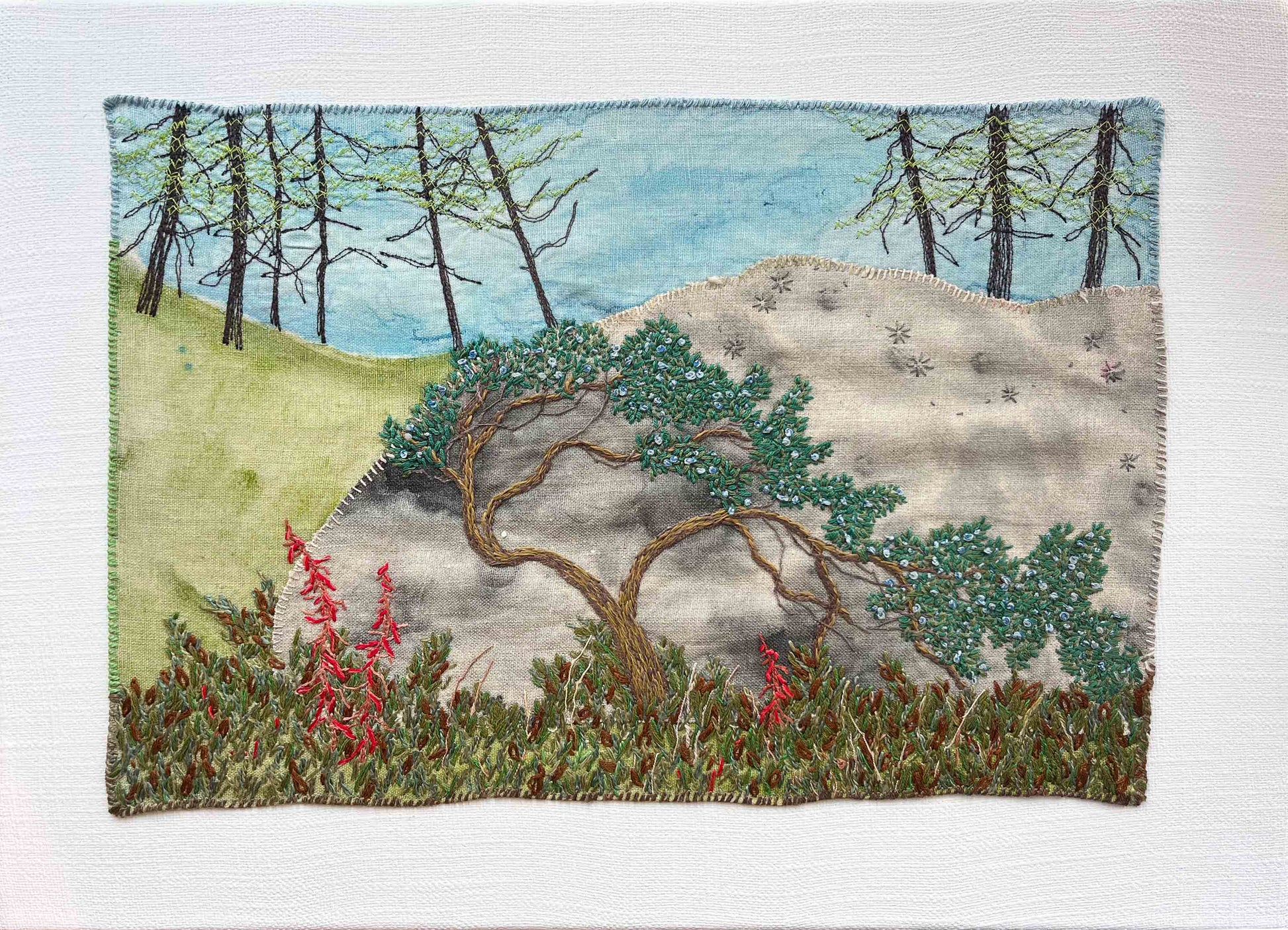 Embroidered wall art: crooked tree on a rocky hill, tall trees in the back, and red flowers in the front.
