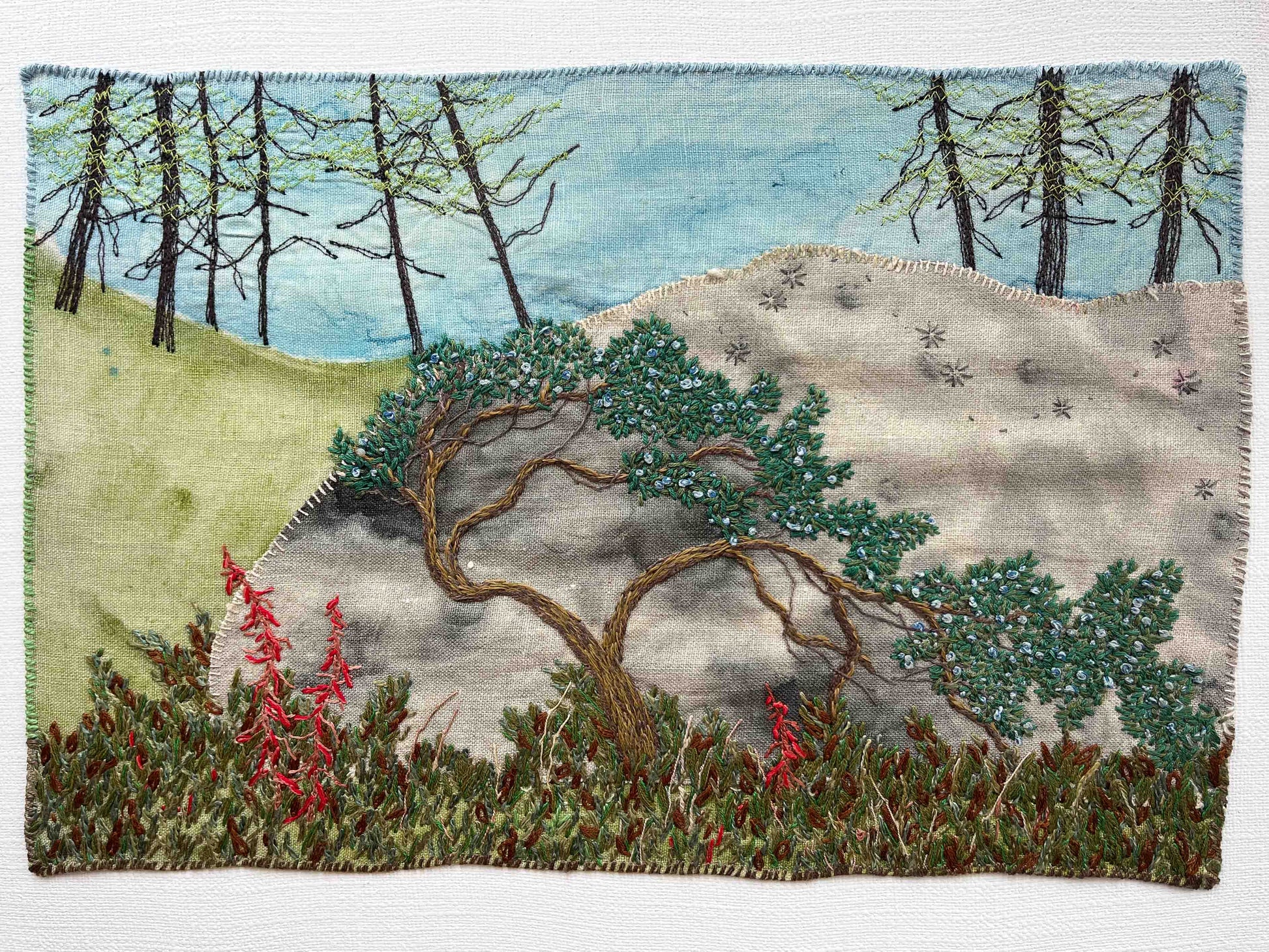 Embroidered wall art: bent tree on a rocky hill, tall trees in the distance, and red flowers in the foreground.