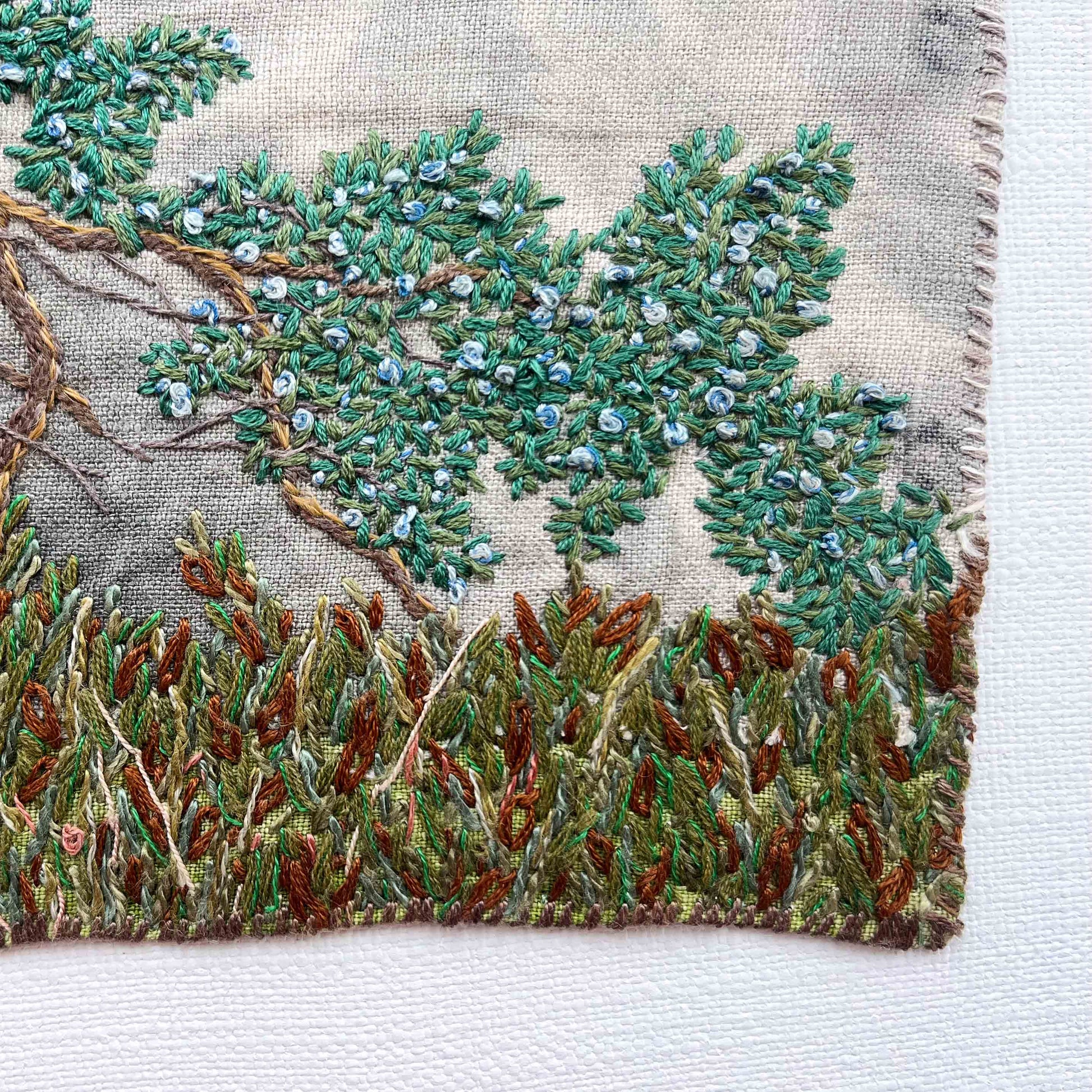 Close-up of embroidered wall art: tree branch with green leaves and blue berries, above textured brown and green foliage.