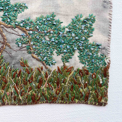 Close-up of embroidered wall art: tree branch with green leaves and blue berries, above textured brown and green foliage.