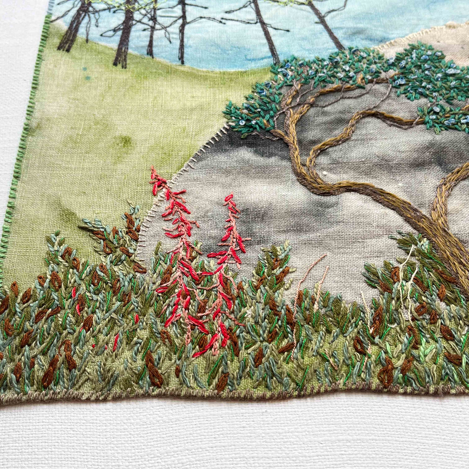 Close-up of embroidered wall art: bent tree, tall trees in the distance, and red flowers in a green foliage patch.