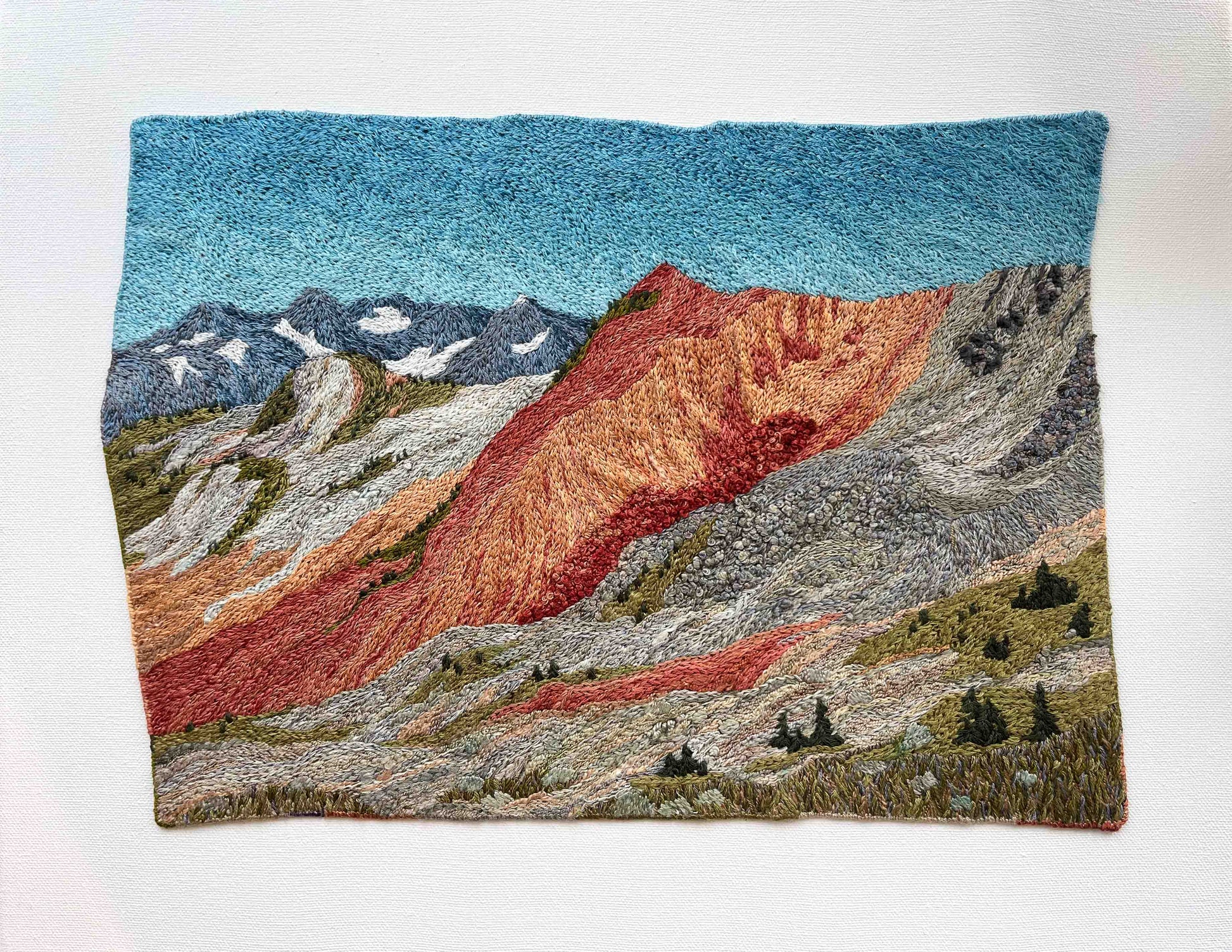 Embroidered wall art: mountain landscape with textured slopes, snow-capped peaks, and small trees.