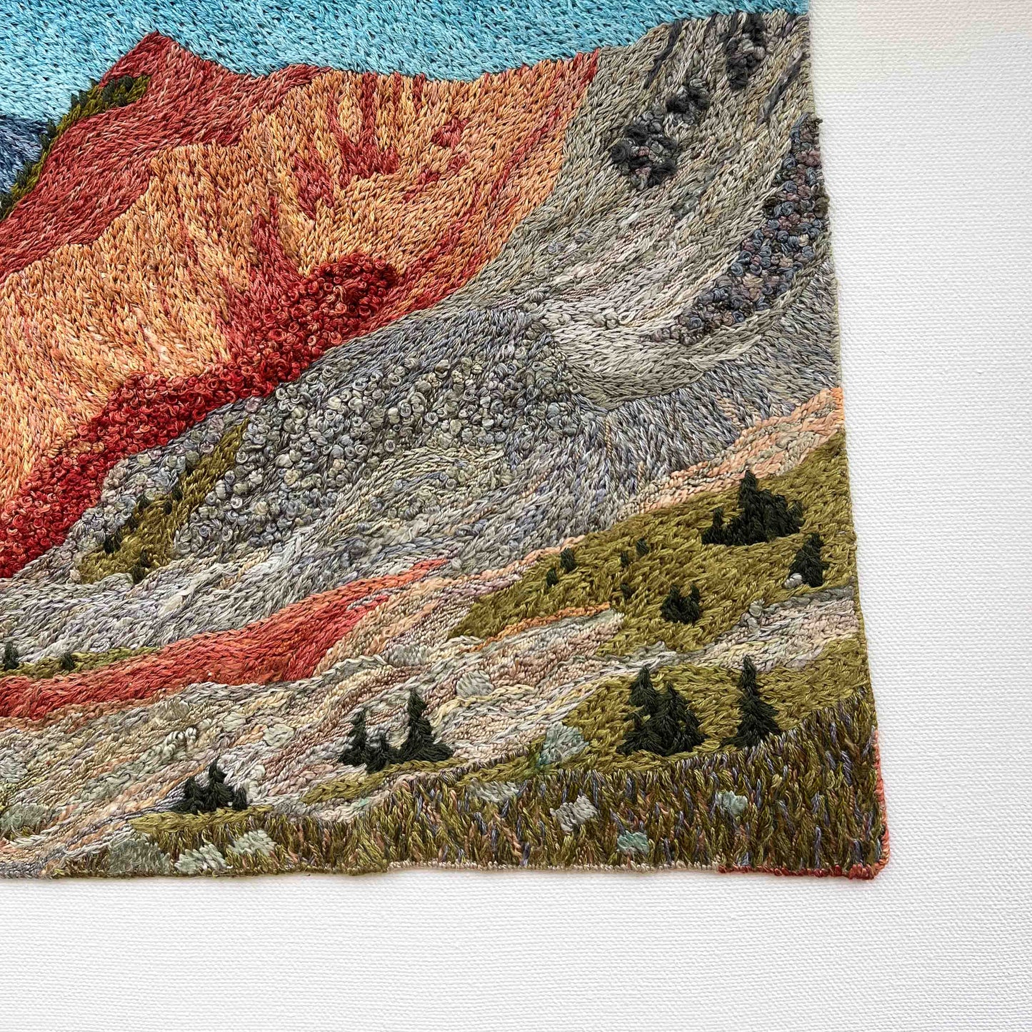 Close-up of embroidered wall art: textured mountain slopes with small trees and detailed stitching.