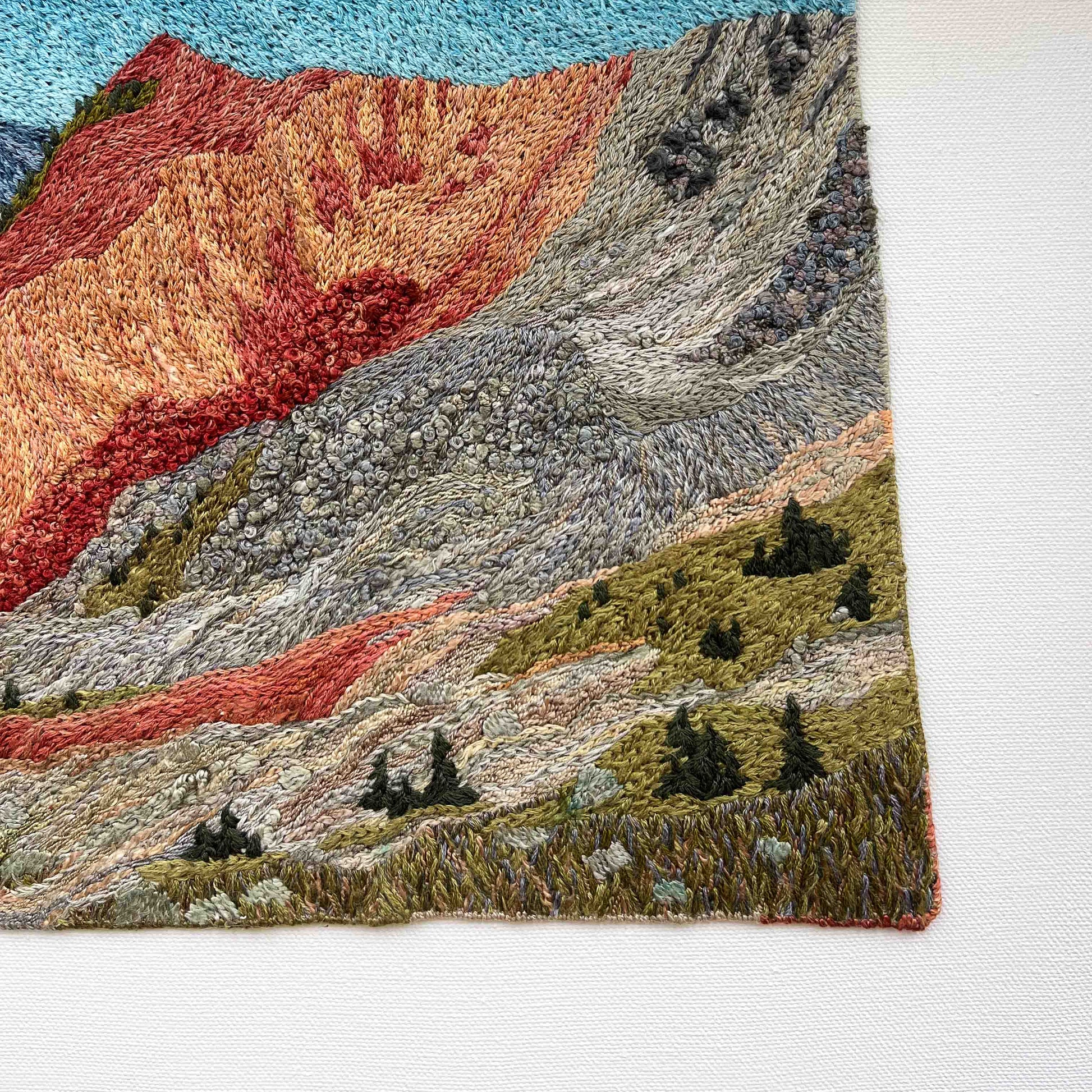 Close-up of embroidered wall art: textured mountain slopes with small trees and detailed stitching.
