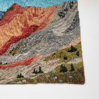 Close-up of embroidered wall art: textured mountain slopes with small trees and detailed stitching.