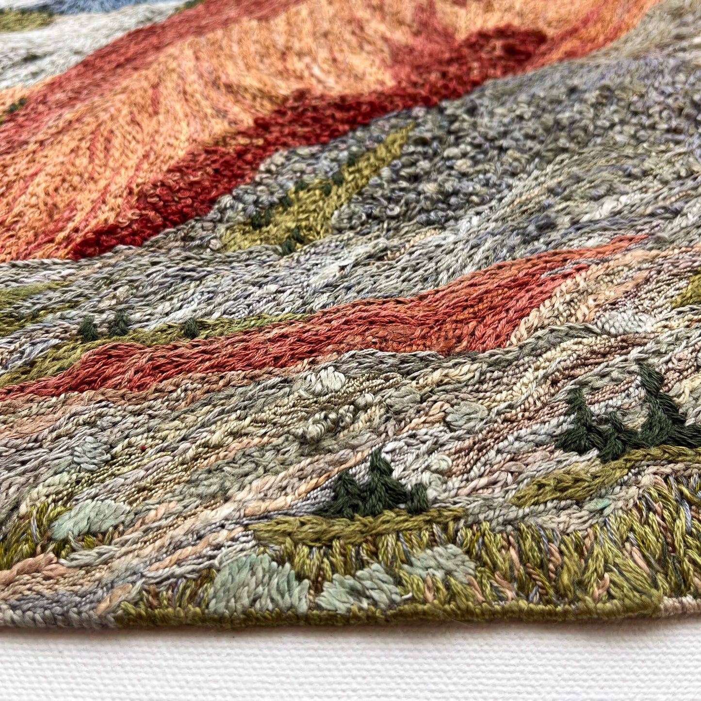 Close-up of embroidered wall art: textured mountain slopes with detailed stitching and small trees.