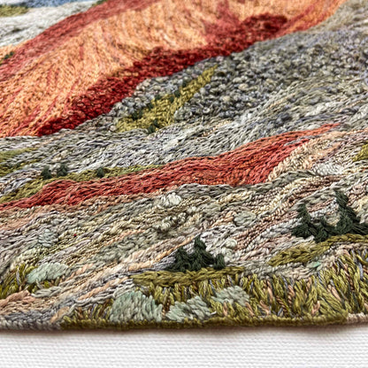 Close-up of embroidered wall art: textured mountain slopes with detailed stitching and small trees.